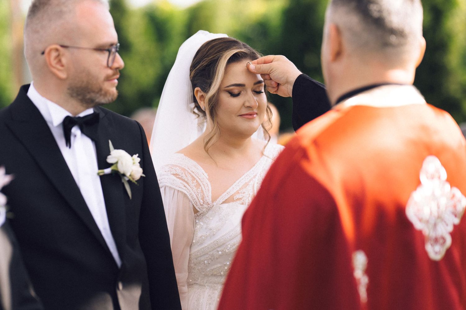 Bride receives a blessing from a priest in red vestments during an outdoor wedding ceremony with groom standing nearby.