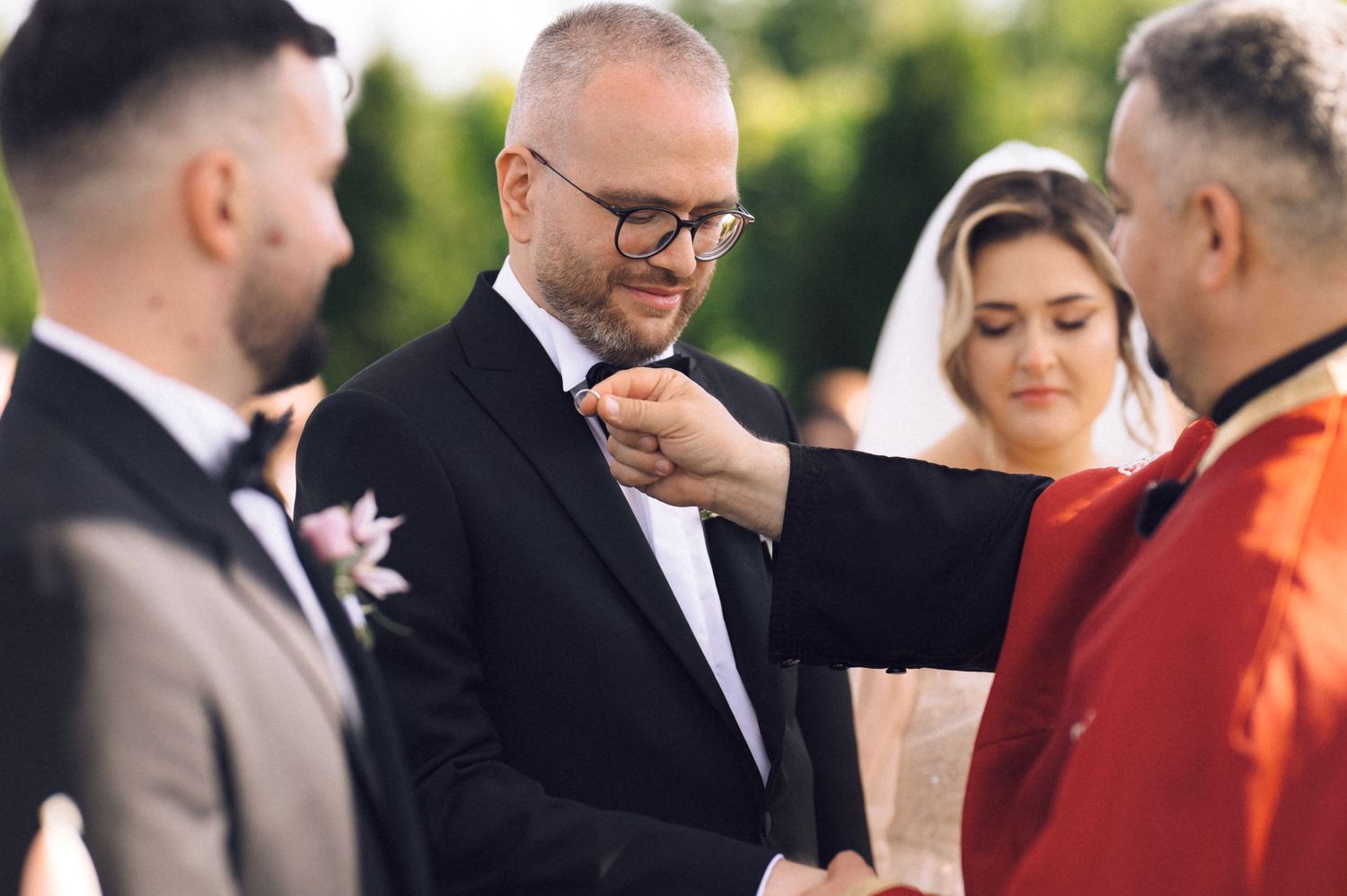A groom in a black tuxedo receives a boutonniere at an outdoor wedding ceremony with guests in the background.