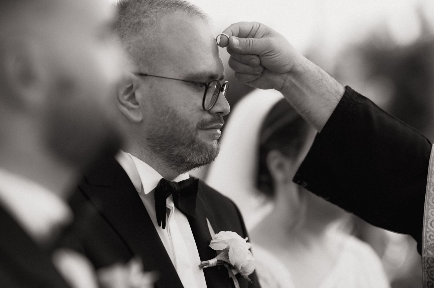 Black and white wedding photo of groom in tuxedo receiving ring during ceremony.