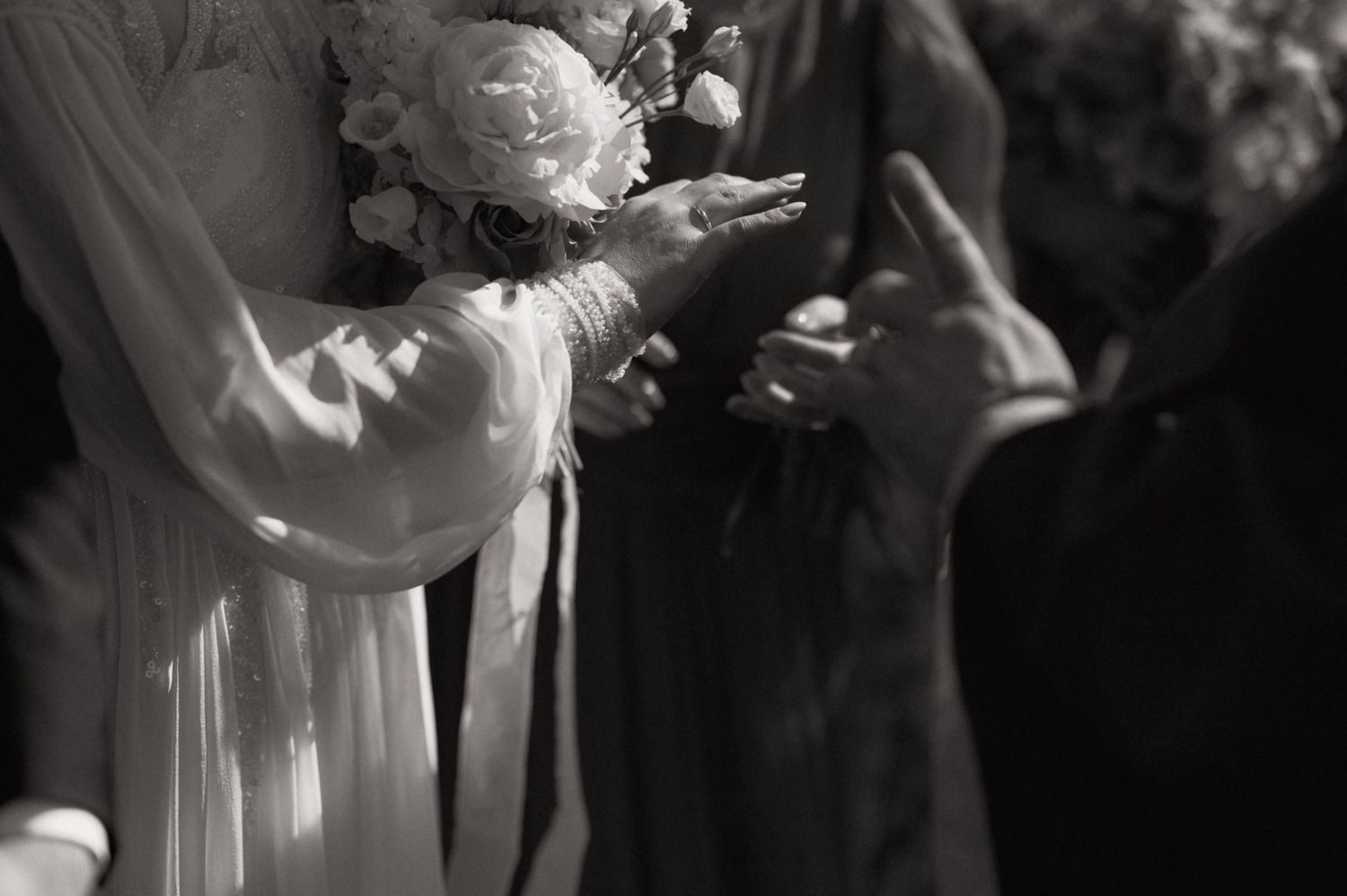 Black and white wedding photo of bride holding white floral bouquet while exchanging handshake during ceremony.