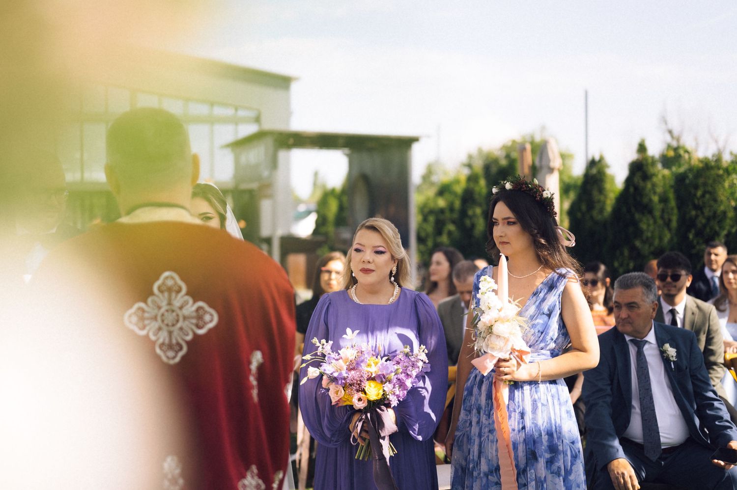 Two women in purple dresses walk down the aisle at an outdoor wedding ceremony holding colorful bouquets.