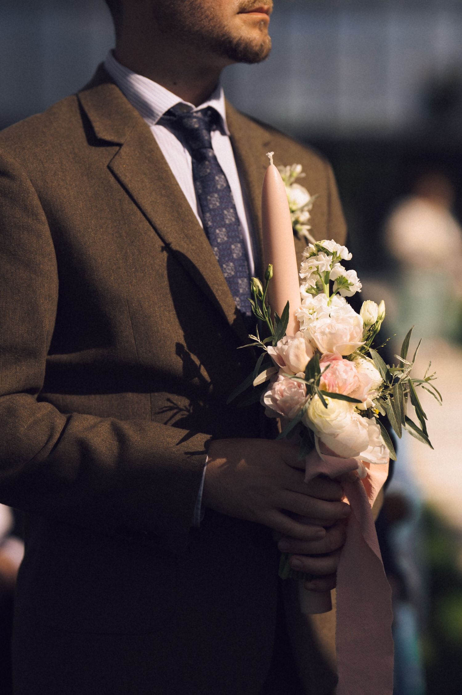 Groom in brown suit holding elegant pink and white floral bouquet with candle at wedding ceremony.