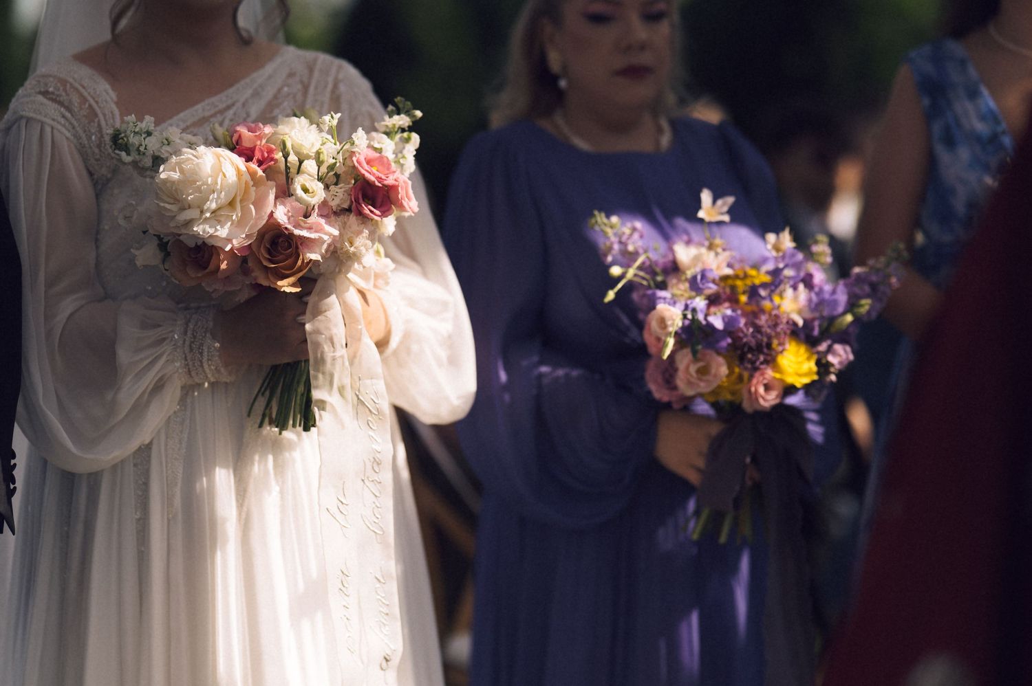 Bride in white gown holding pink rose bouquet beside bridesmaid in blue dress with purple wildflower bouquet at wedding.