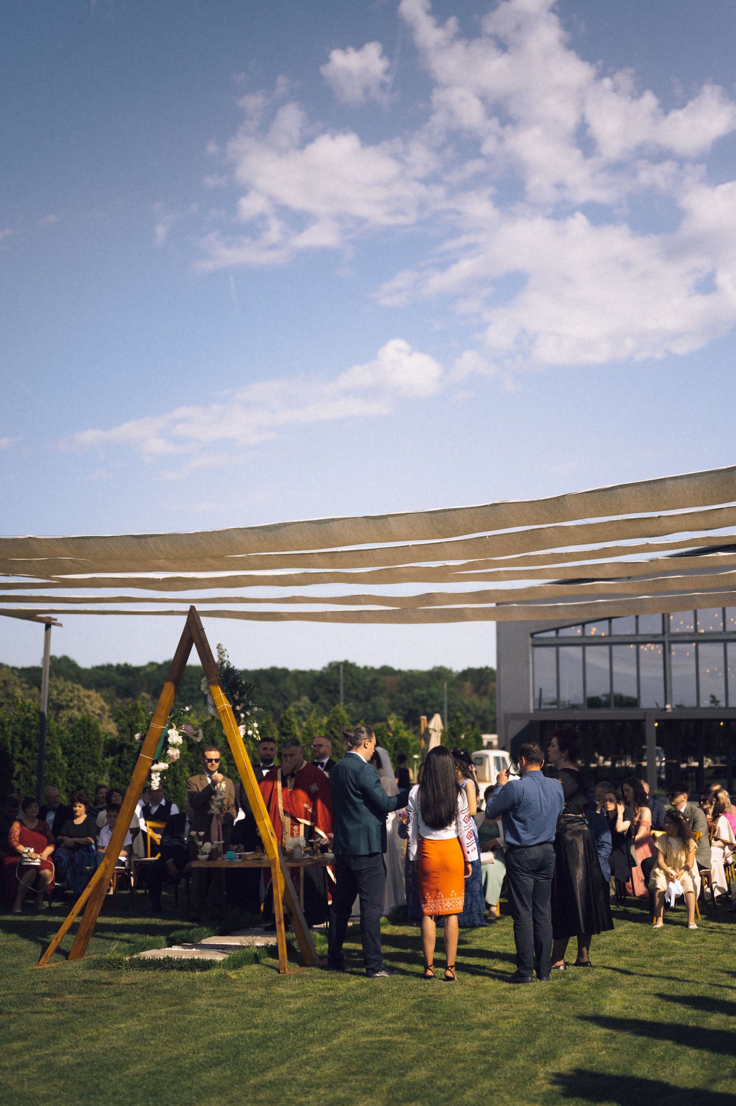 Outdoor wedding reception on green lawn with guests mingling under fabric canopy, wooden arch, and blue sky.
