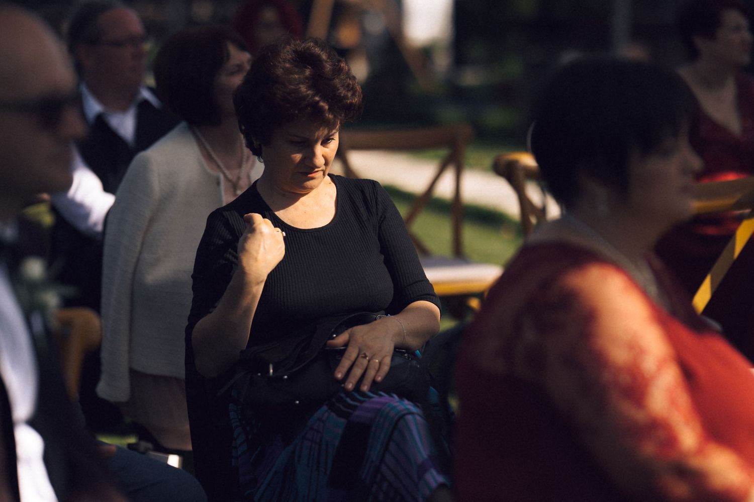 Woman in black top sitting outdoors at an evening event, surrounded by other attendees in warm ambient light.