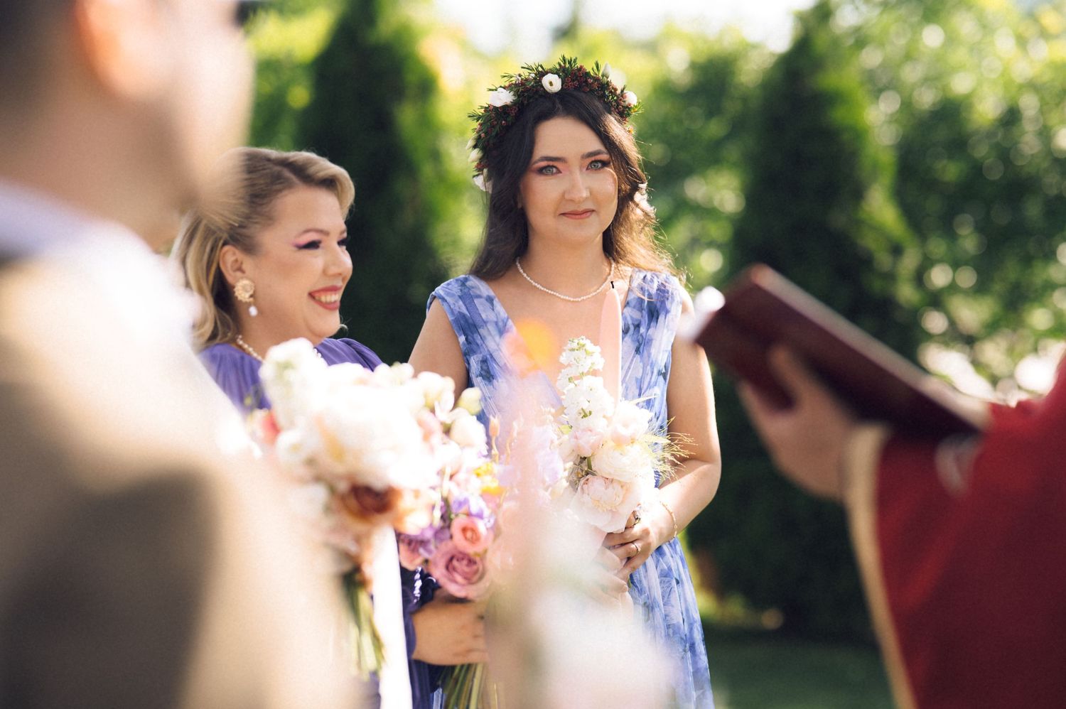 Two women in blue floral dresses holding bouquets smile during an outdoor wedding ceremony with greenery background.