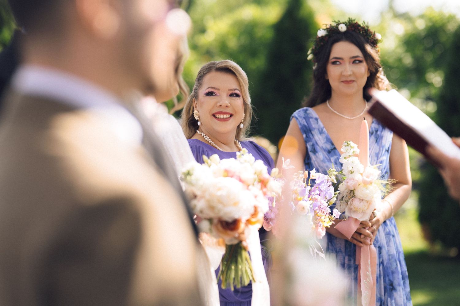 Two smiling bridesmaids in blue and purple dresses holding bouquets at an outdoor wedding ceremony.
