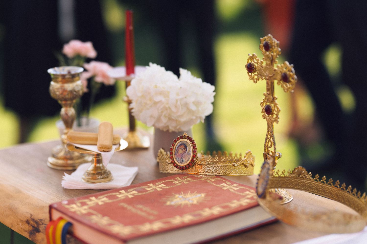Orthodox wedding ceremony items including a gold cross, crowns, Bible, chalice, and white flowers on a wooden table.
