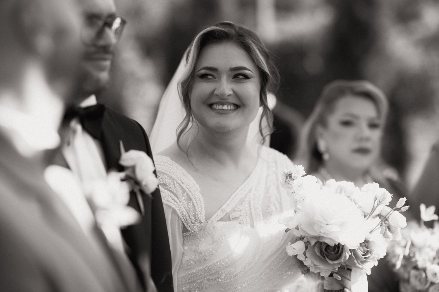 Black and white photo of a smiling bride in a veil holding a bouquet at her wedding ceremony.