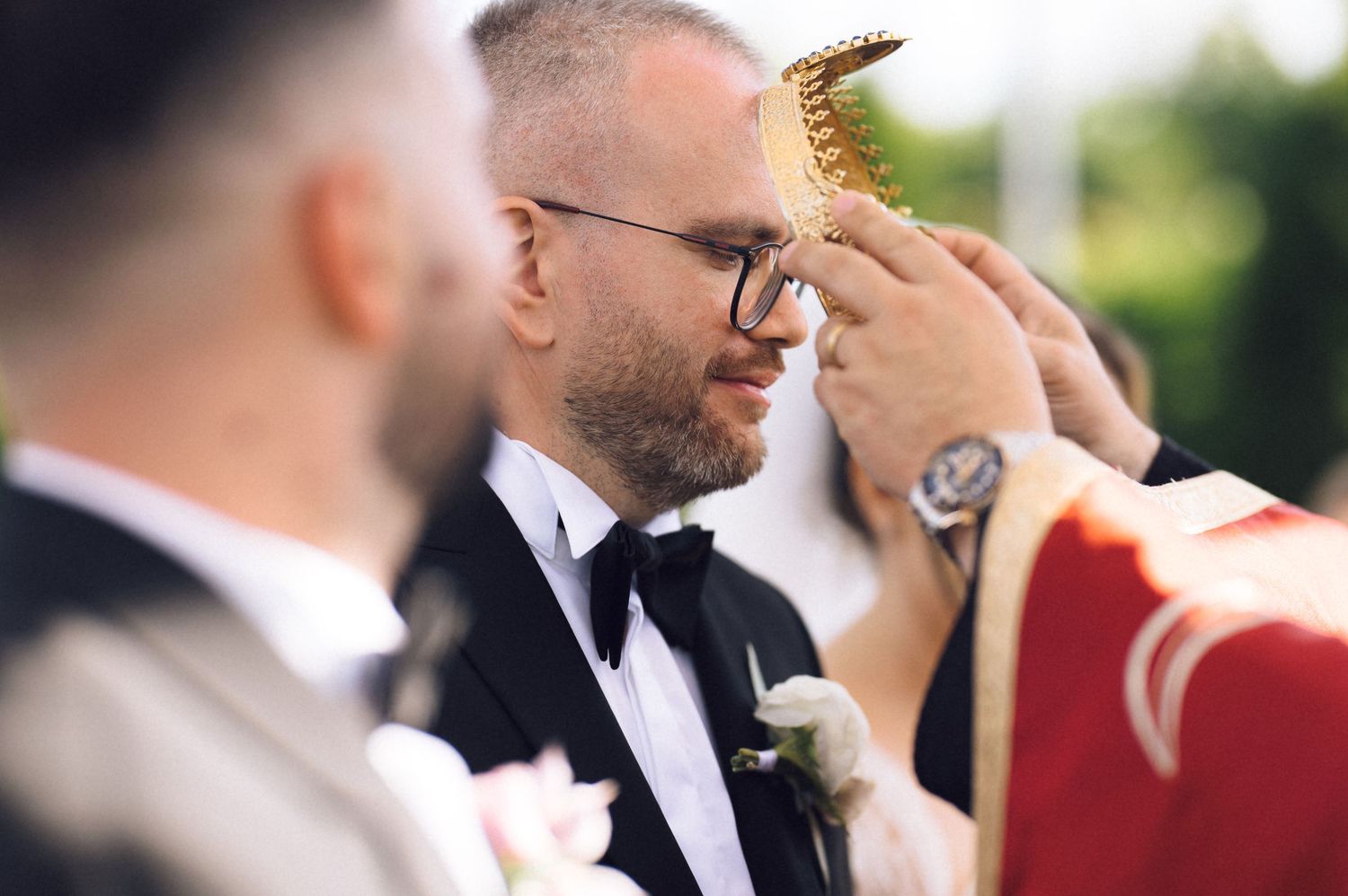 Groom in tuxedo receives anointing during wedding ceremony outdoors.