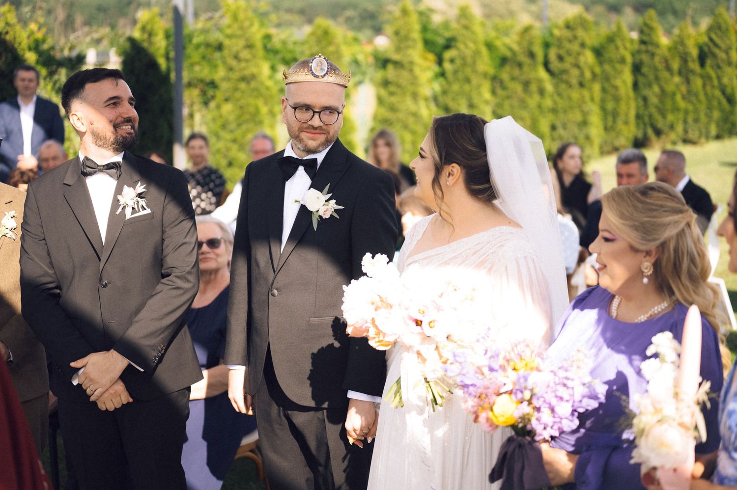 Outdoor wedding ceremony with groom in gray suit, officiant, bride in white gown holding flowers, and guests gathered in garden.