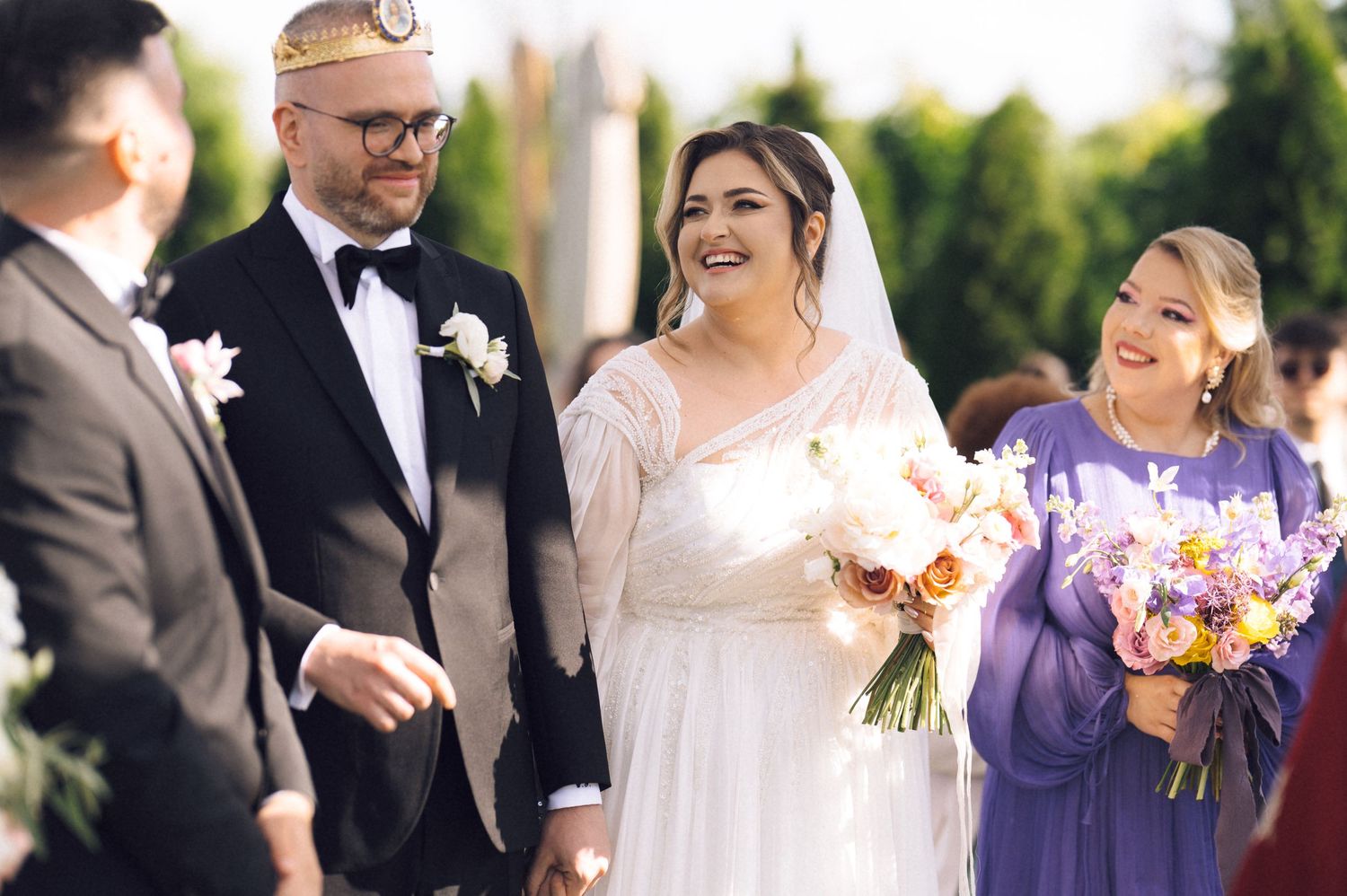Happy bride and groom walk down the aisle at outdoor wedding ceremony with smiling bridesmaid in purple dress.