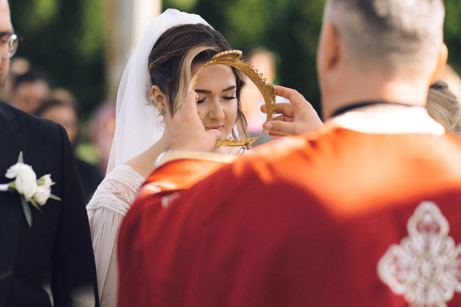 Bride receiving a blessing from a priest in red vestments during an outdoor Orthodox wedding ceremony.
