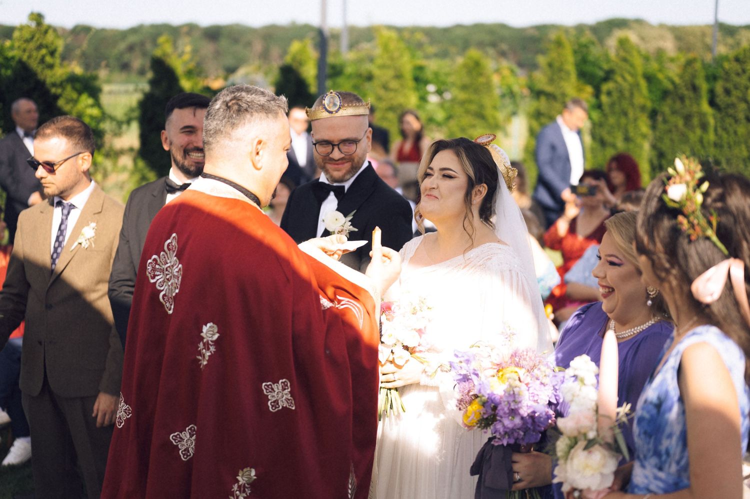 Outdoor wedding ceremony with bride holding bouquet, groom, and officiant in red robe, surrounded by guests.