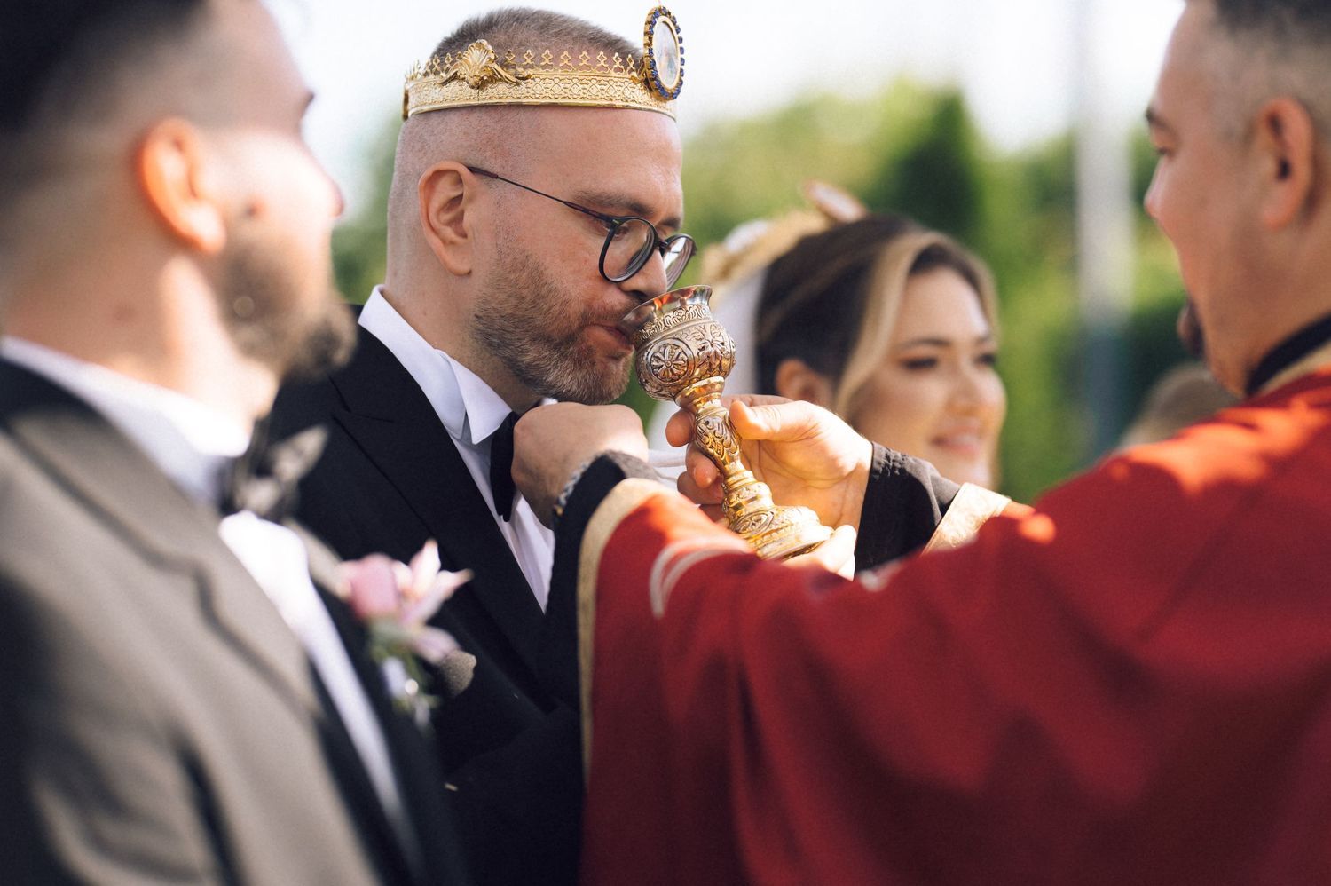 Groom wearing crown kisses chalice during Orthodox wedding ceremony outdoors.