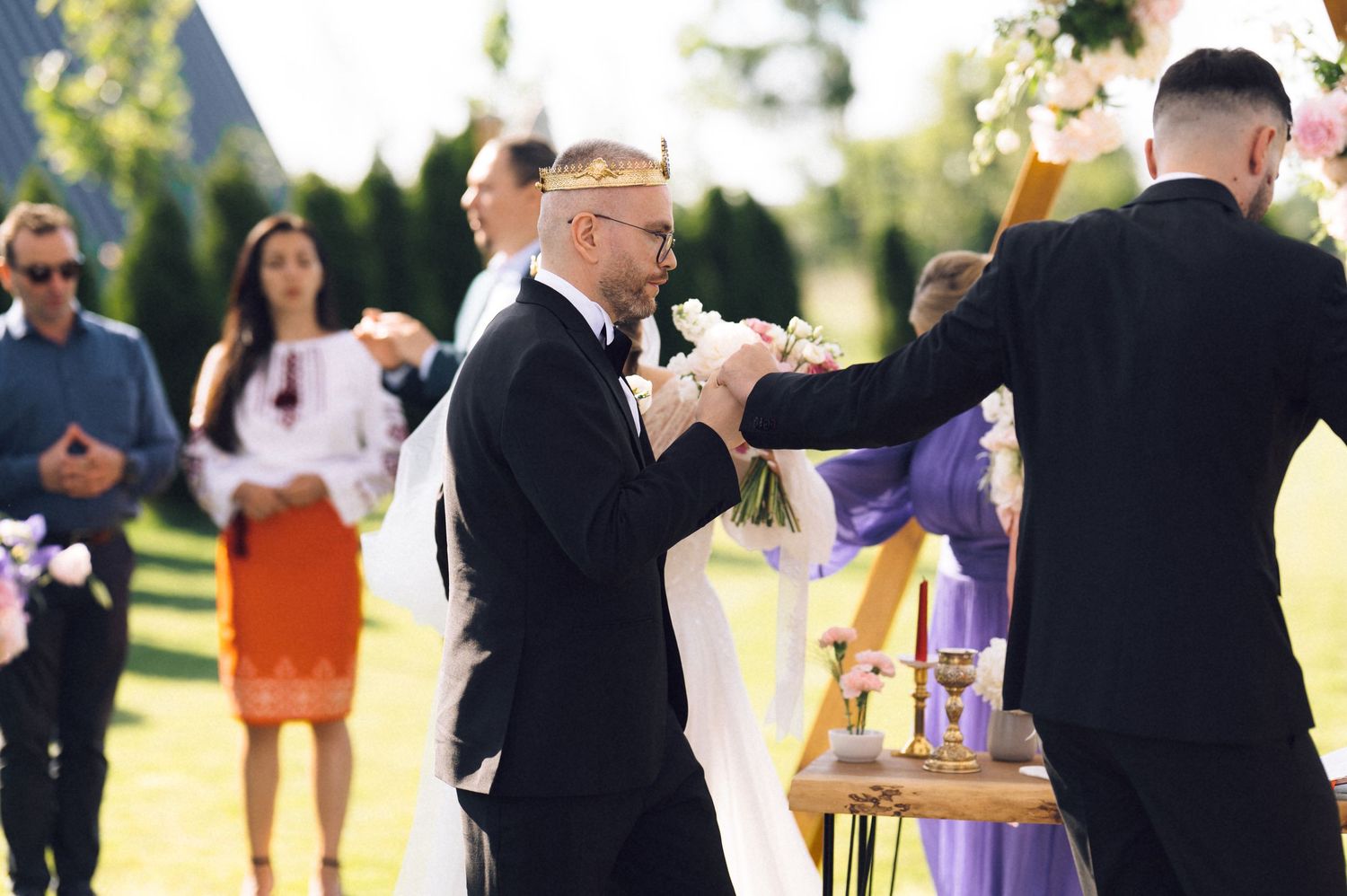 Groom shaking hands with groomsman at outdoor wedding ceremony with guests in background.