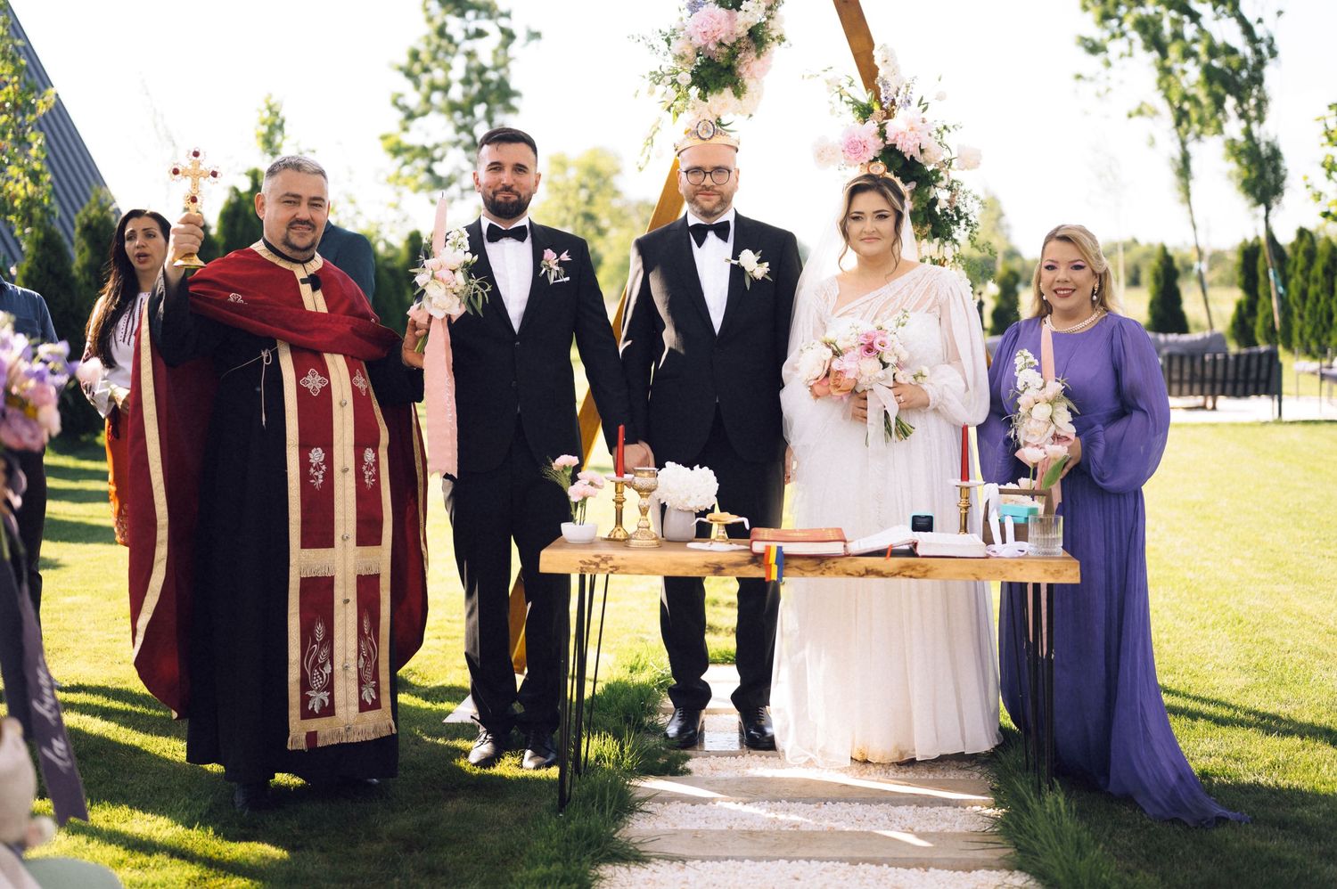 Orthodox priest officiates outdoor wedding ceremony with bride, groom, and guests.