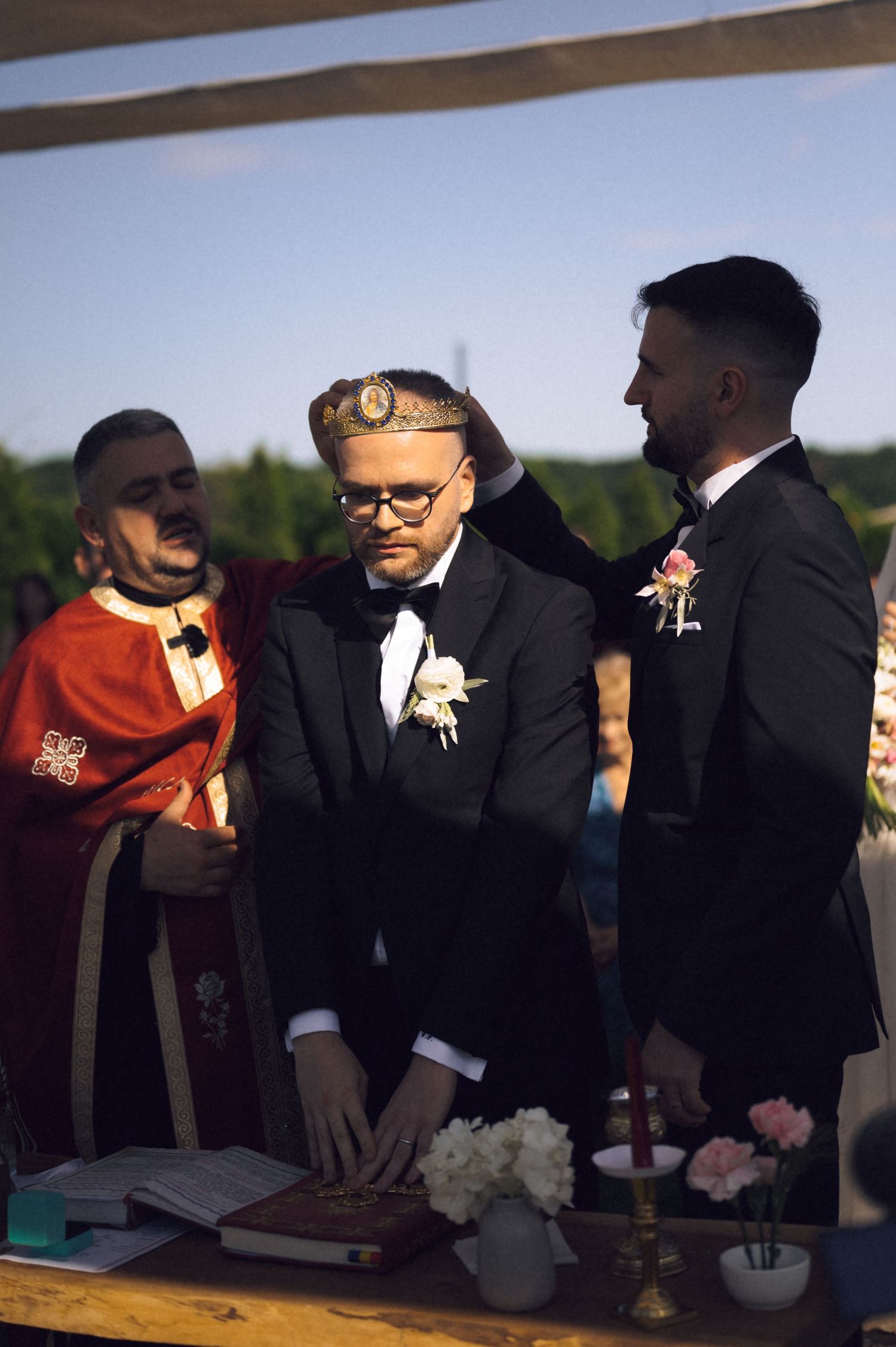Two grooms in tuxedos share a wedding ceremony moment with an officiant in a red robe at an outdoor venue.