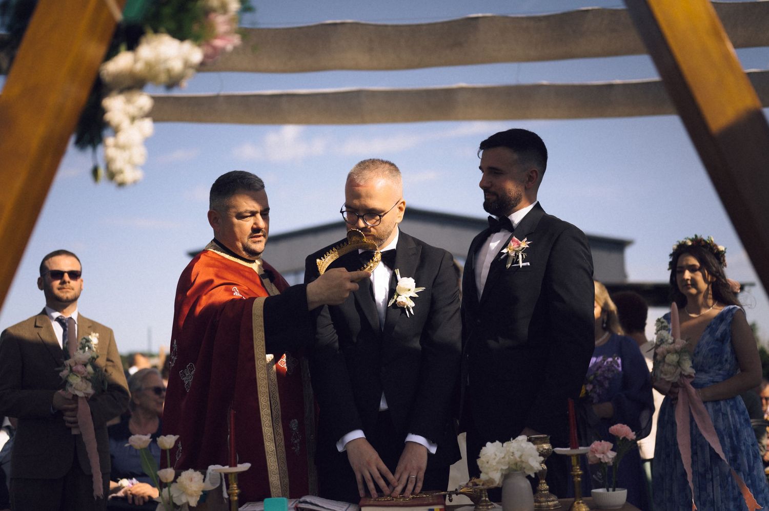 Three men in formal attire stand at an outdoor wedding ceremony under a decorated arch.