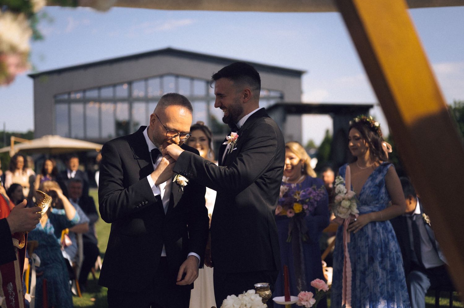 Two grooms share an emotional moment during their outdoor wedding ceremony, one wiping tears while guests watch.
