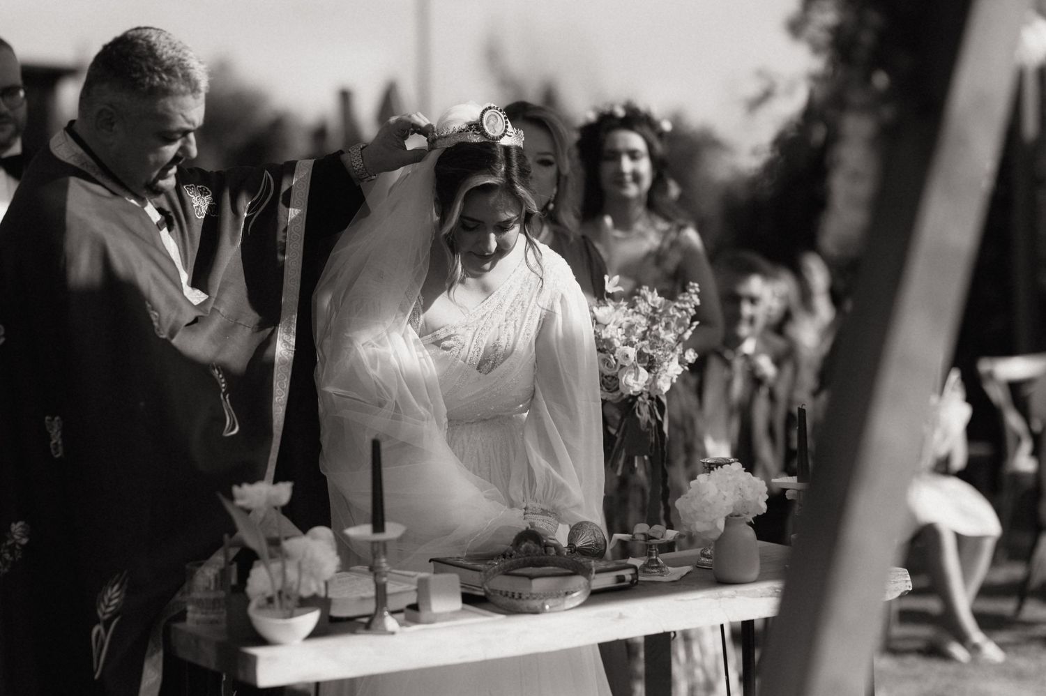 Black and white photo of a bride in a white veil leaning over a table during an outdoor wedding ceremony.