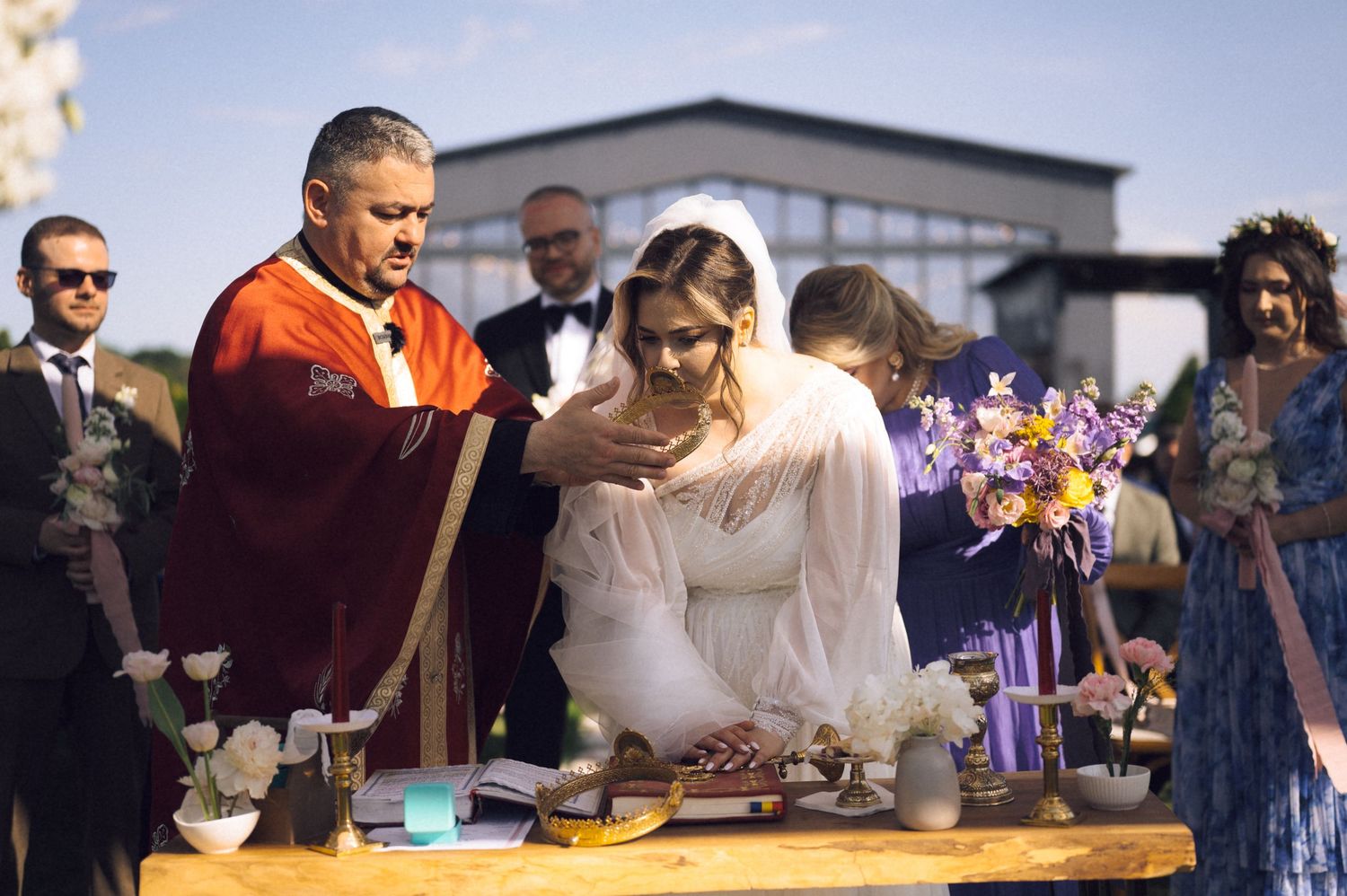 Bride in white gown participates in outdoor wedding ceremony with officiant in red robe and guests in formal attire.