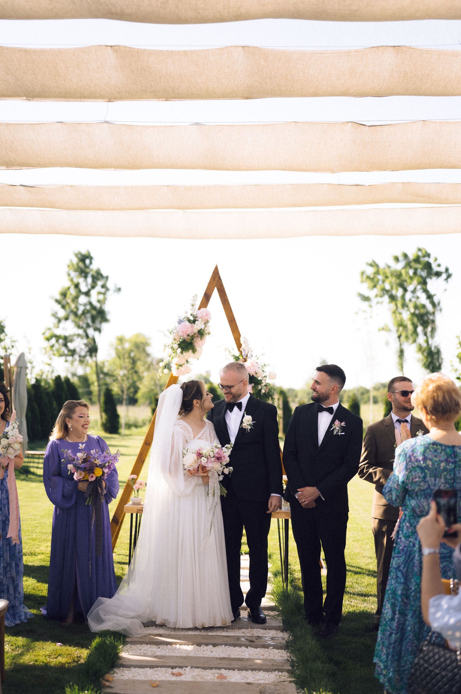 Bride and groom stand at a golden triangle arch adorned with pink flowers during an outdoor wedding ceremony.