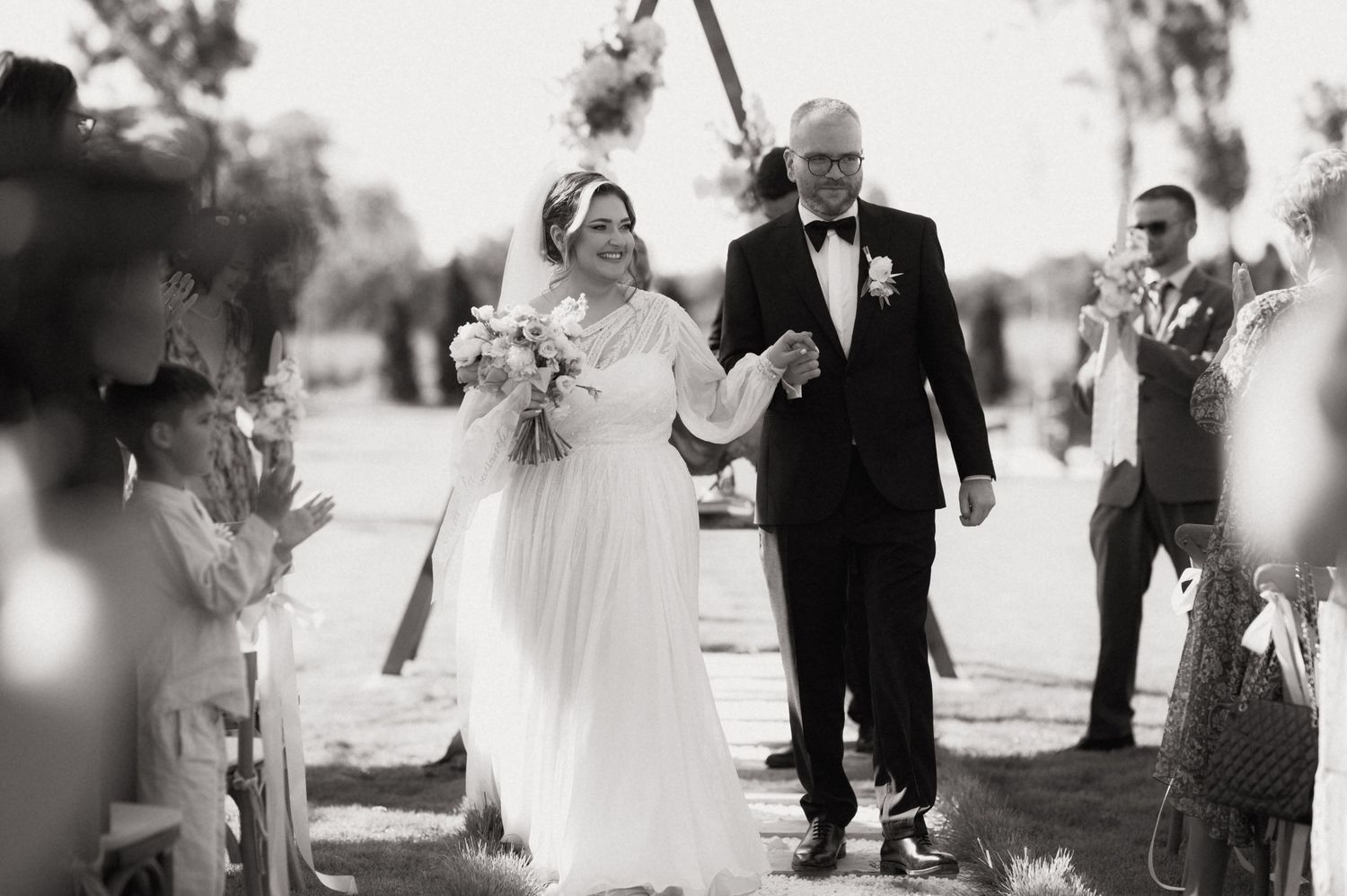 Black and white photo of a smiling bride and groom walking down the aisle at an outdoor wedding ceremony.