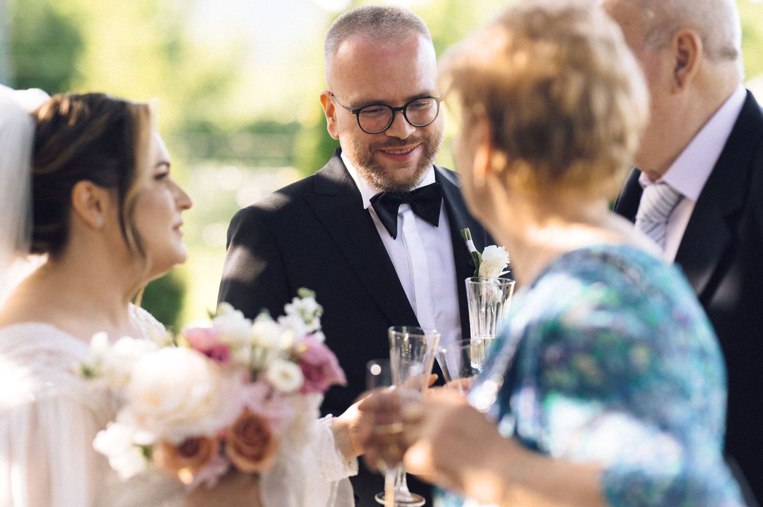 Bride and groom celebrating with guests at outdoor wedding reception, holding champagne glasses and floral bouquet.