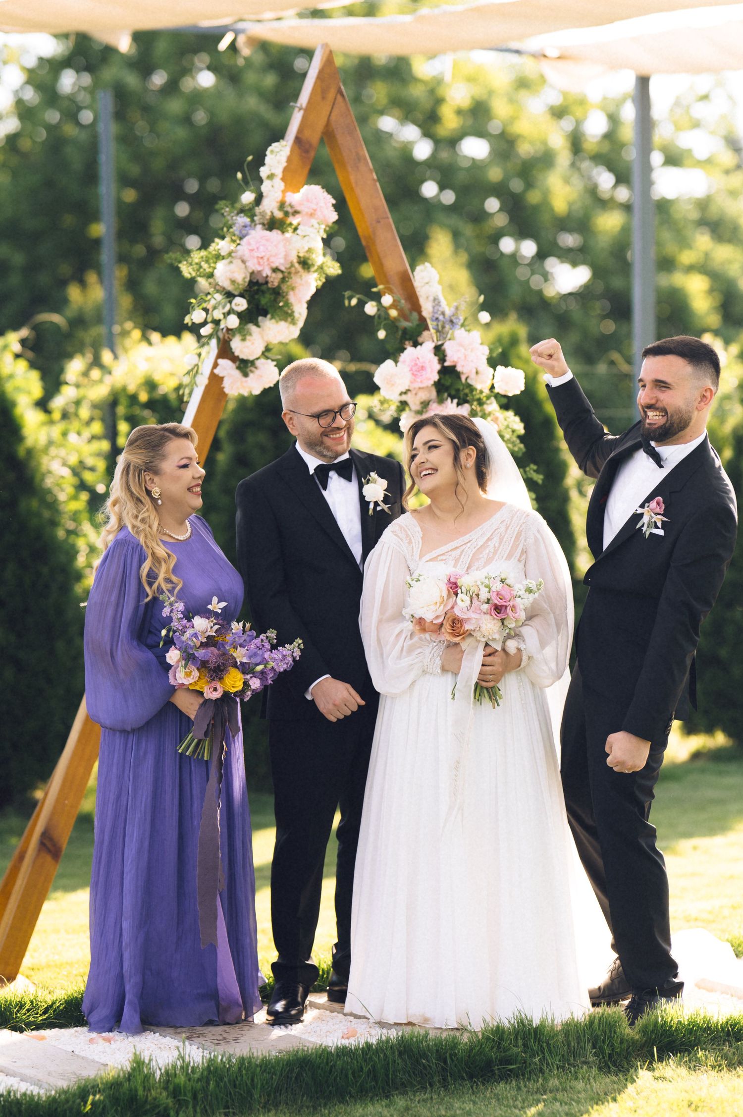Joyful wedding ceremony with bride in white gown, groom celebrating, officiant, and bridesmaid in purple dress outdoors.