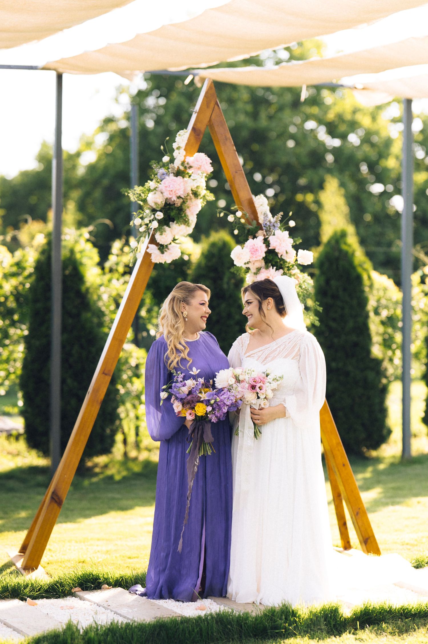 Two brides stand under a wooden triangular arch adorned with pink flowers, holding bouquets at an outdoor wedding ceremony.