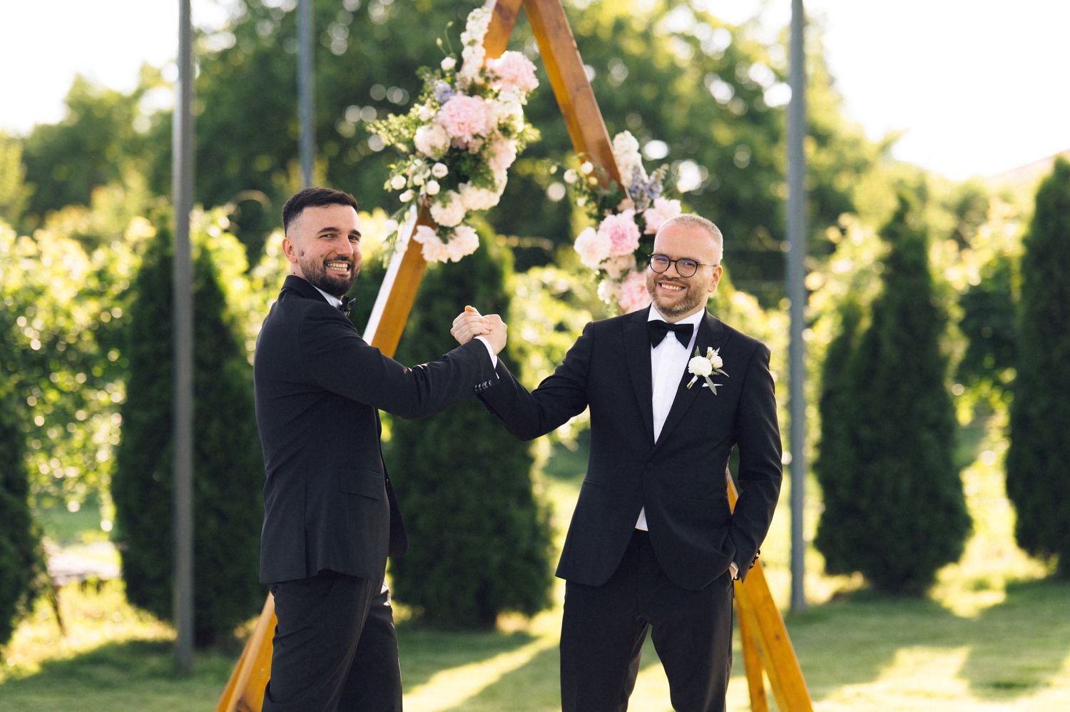 Two grooms in black tuxedos share a joyful moment at an outdoor wedding ceremony with a floral arch.