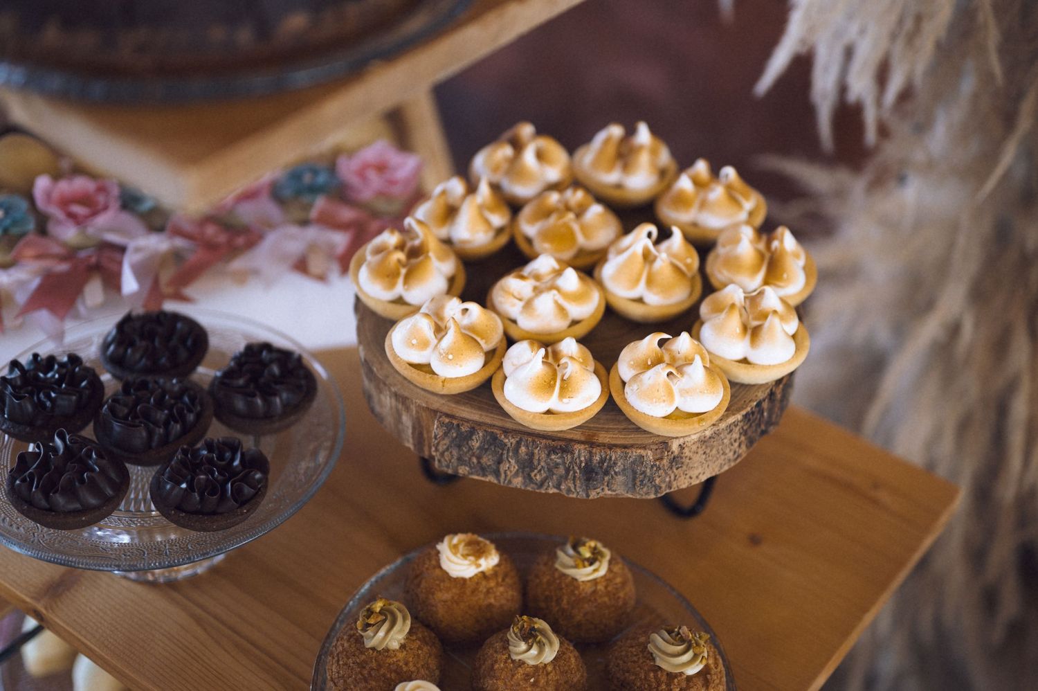 Elegant dessert table featuring gold-topped meringue cupcakes on a rustic wood stand, with dark chocolate treats on a glass plate.