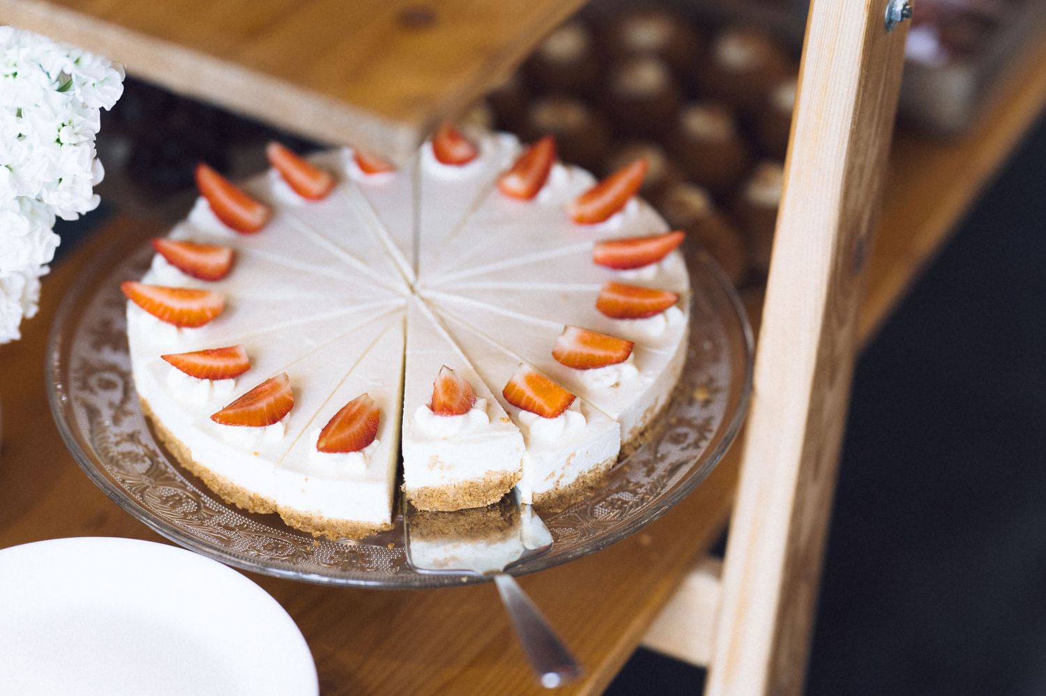 Sliced cheesecake topped with fresh strawberries on a decorative glass cake stand at a dessert table.