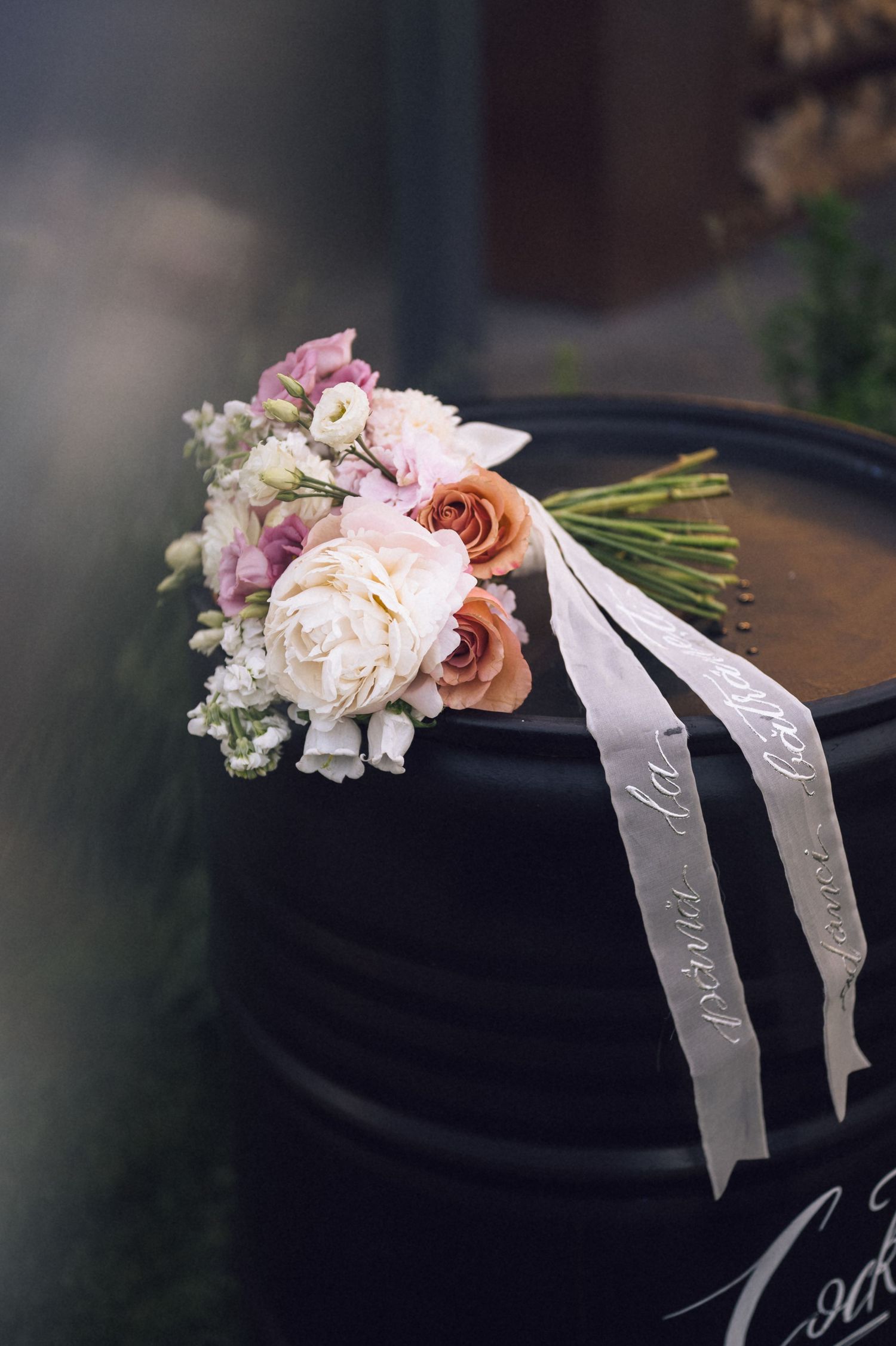 Elegant wedding bouquet with white peonies and peach roses, tied with a calligraphy ribbon, resting on a black barrel.