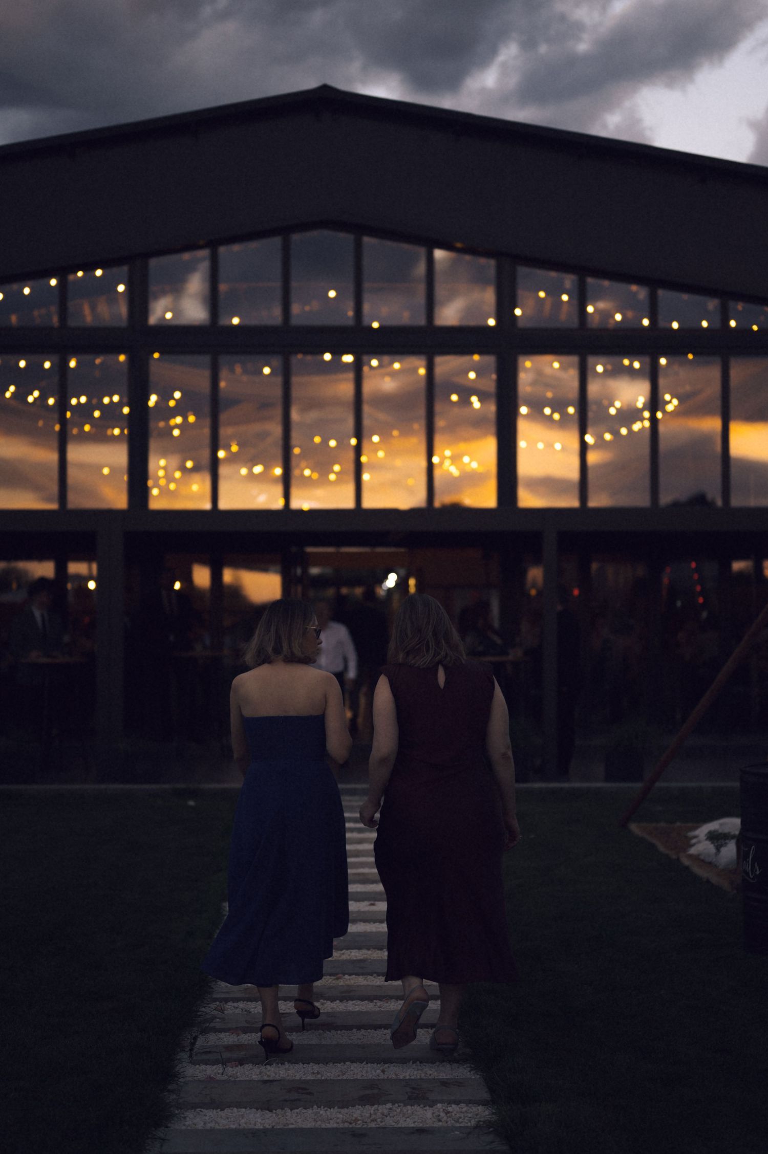 Two women in elegant dresses walk toward a warmly lit venue at dusk, with string lights glowing inside.