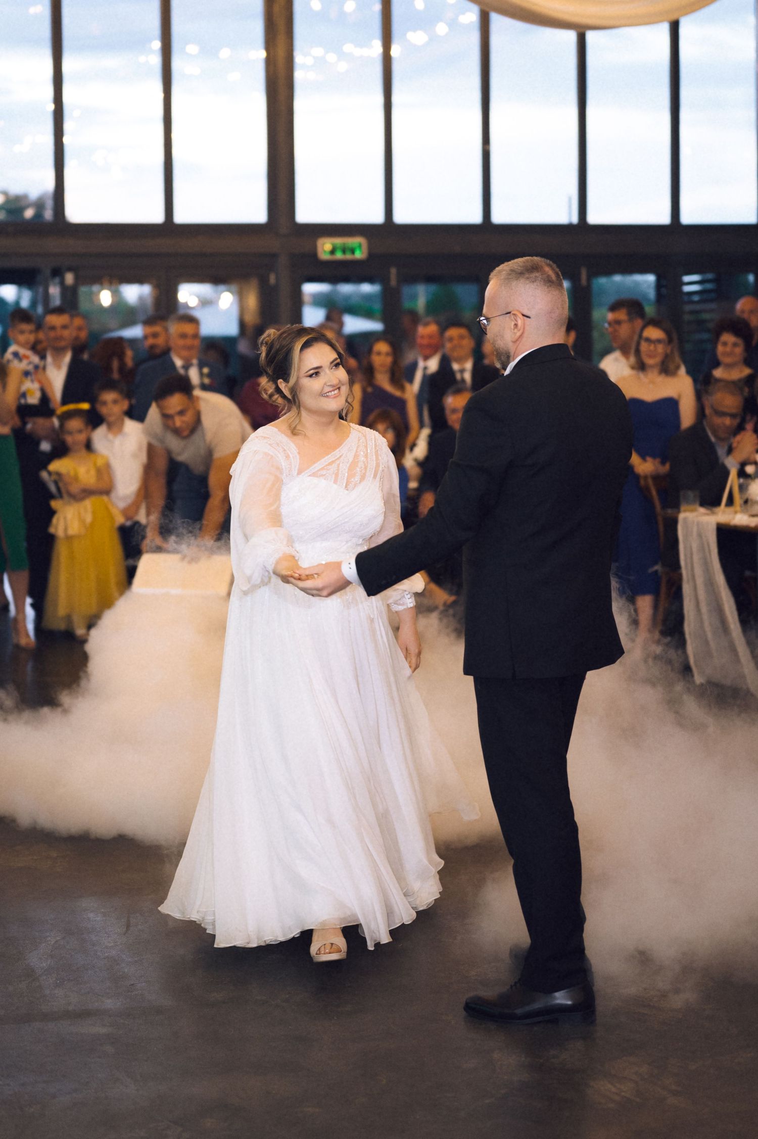 Bride and groom share their first dance surrounded by dry ice fog at a wedding reception with guests watching.