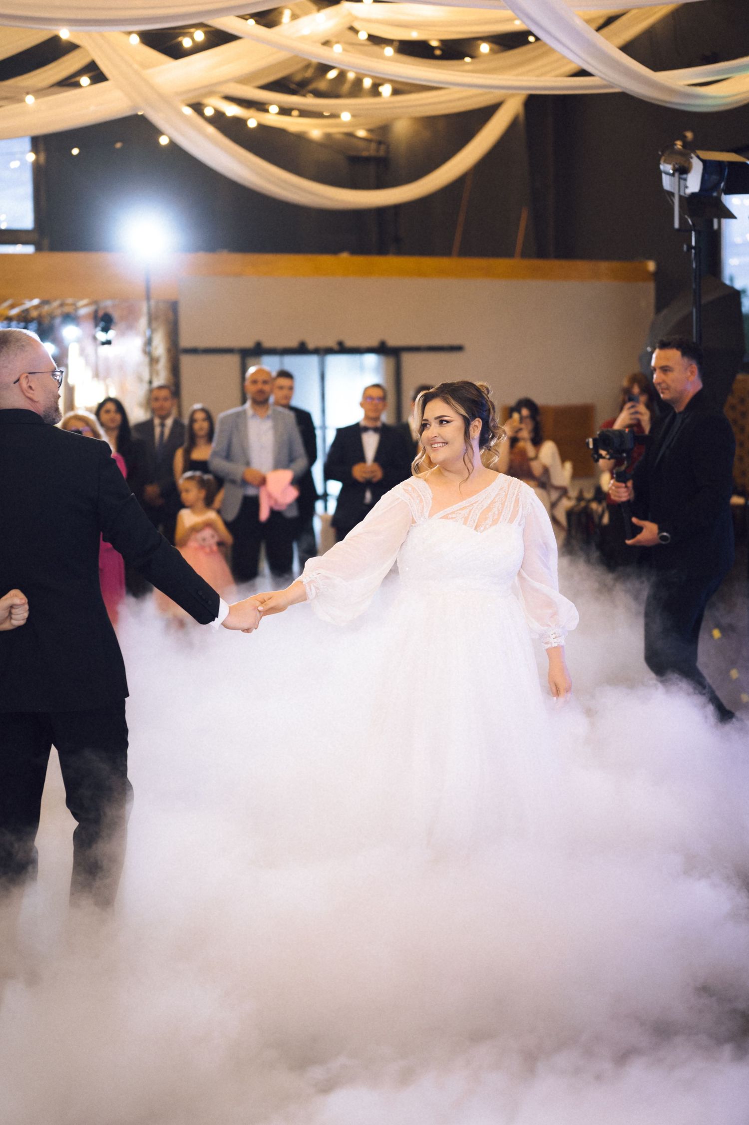 Bride in a white gown dances with groom on a fog-covered dance floor at a romantic wedding reception.