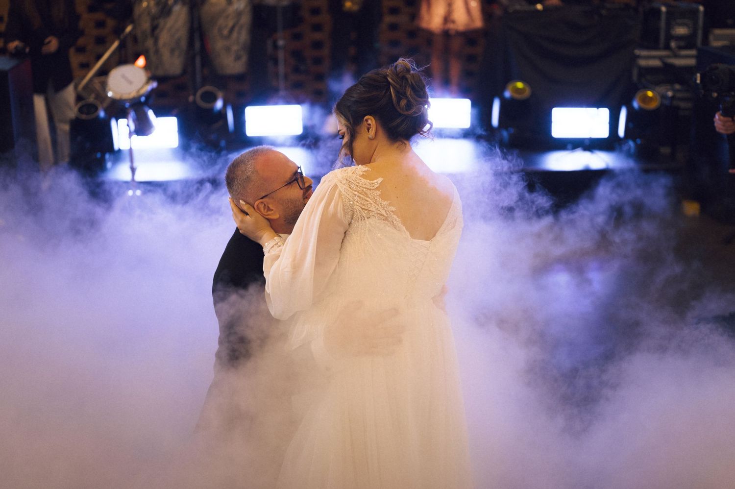 Bride and groom share first dance surrounded by dramatic fog on a dimly lit wedding reception dance floor.