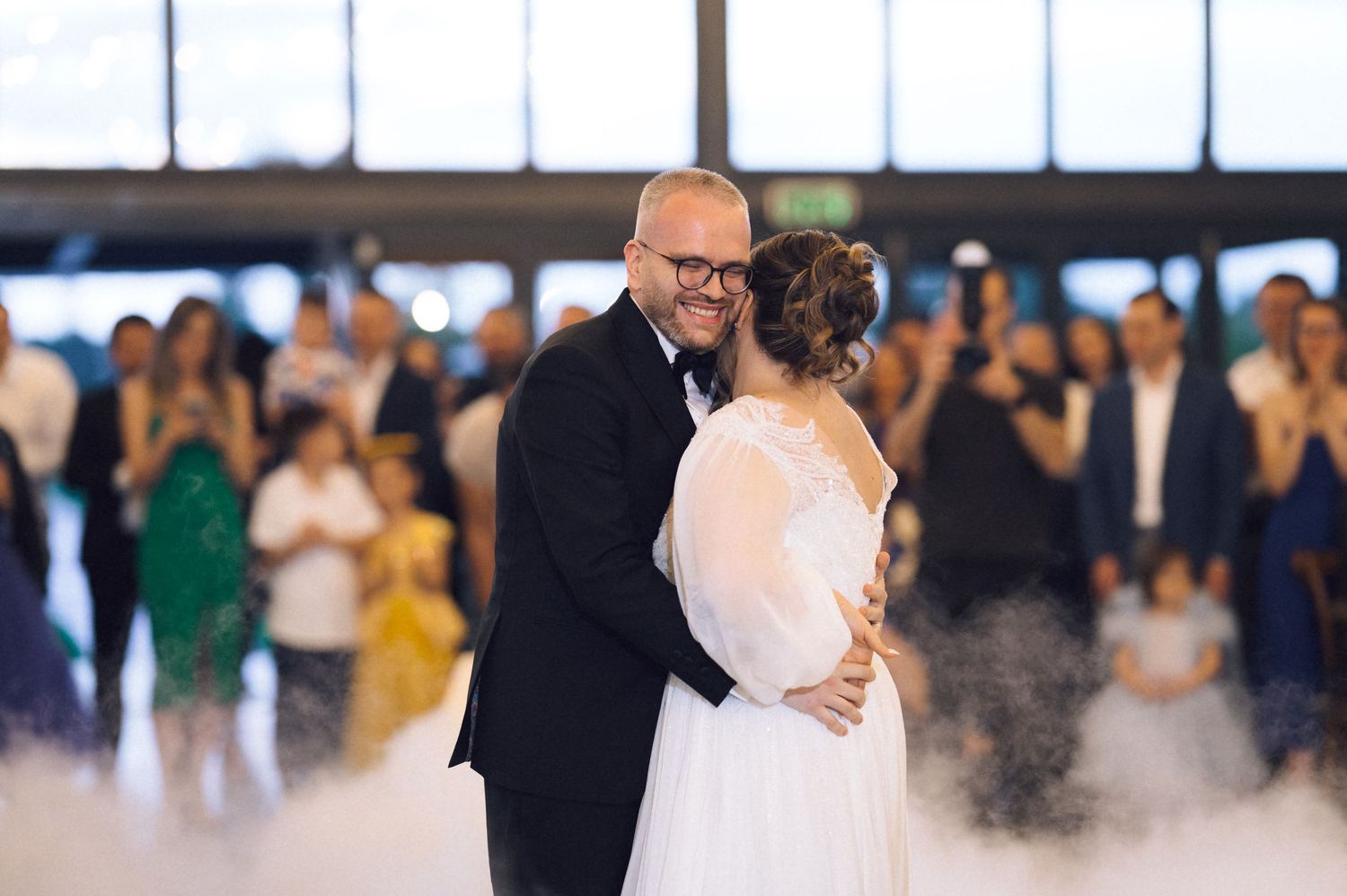 Bride and groom share their first dance surrounded by guests at an elegant wedding reception with atmospheric fog effects.