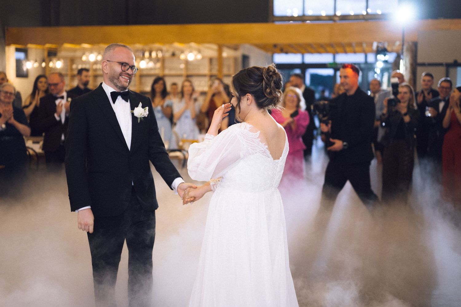 Bride and groom share first dance on fog-covered floor at elegant wedding reception with guests watching.