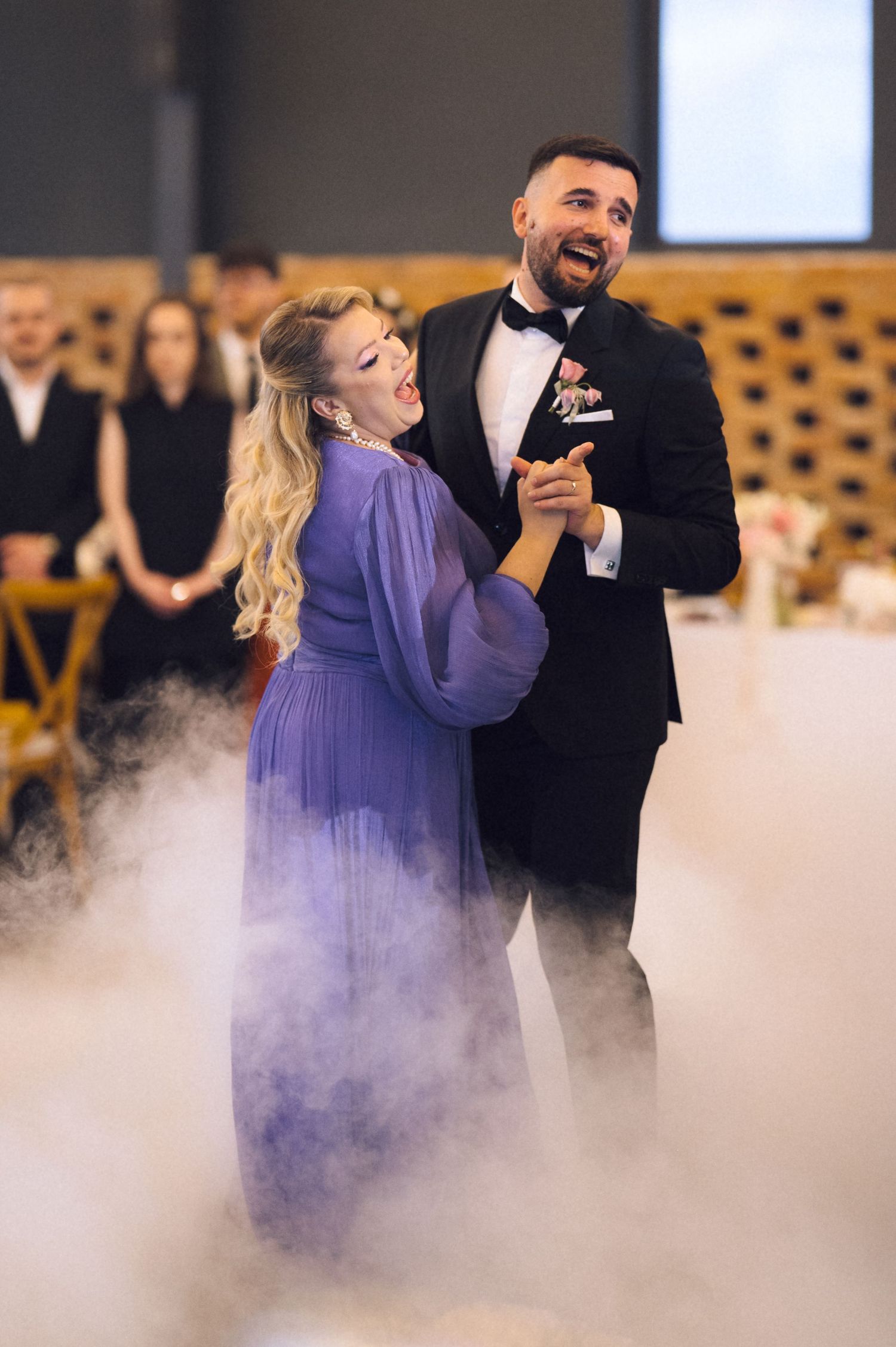 Couple sharing first dance at wedding reception surrounded by dramatic fog effect, woman in purple gown, man in black tuxedo.