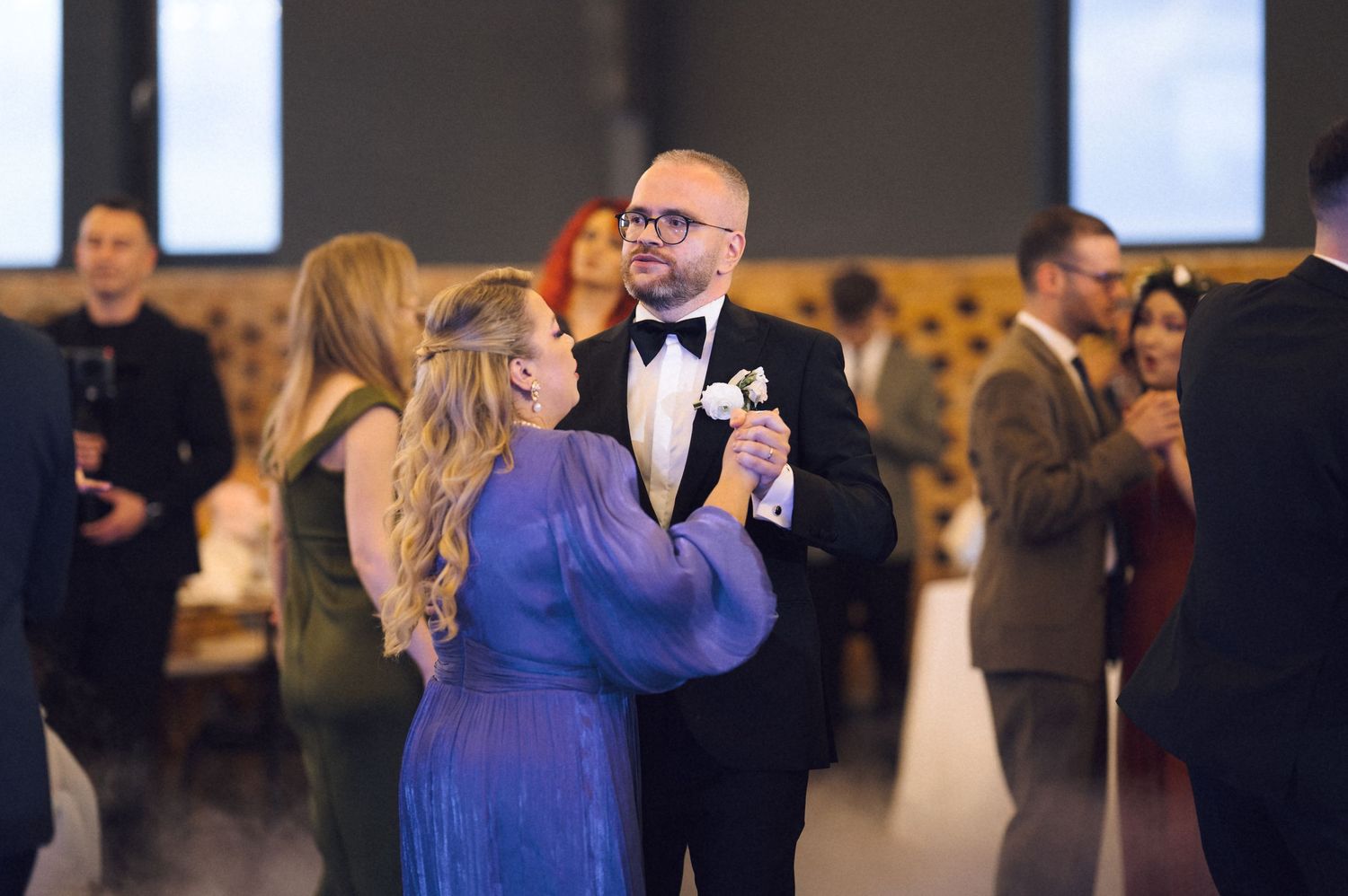 A groom in a black tuxedo shares a dance with a woman in a blue dress at an elegant wedding reception.