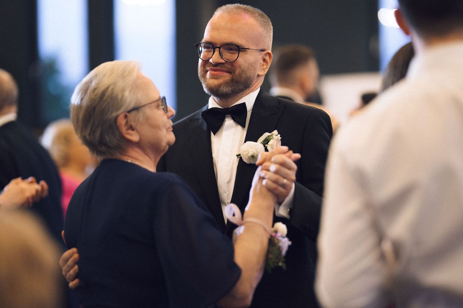 Groom in tuxedo sharing a dance with elderly woman at wedding reception.