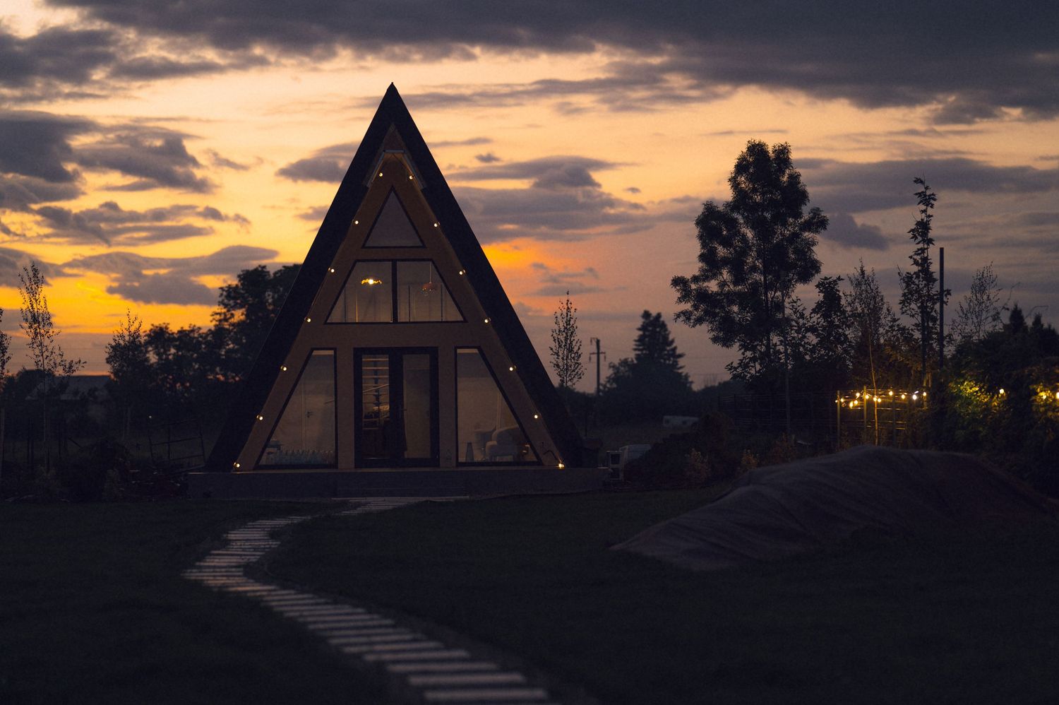 A-frame cabin with string lights glows at dusk under dramatic orange and purple sunset sky surrounded by trees.
