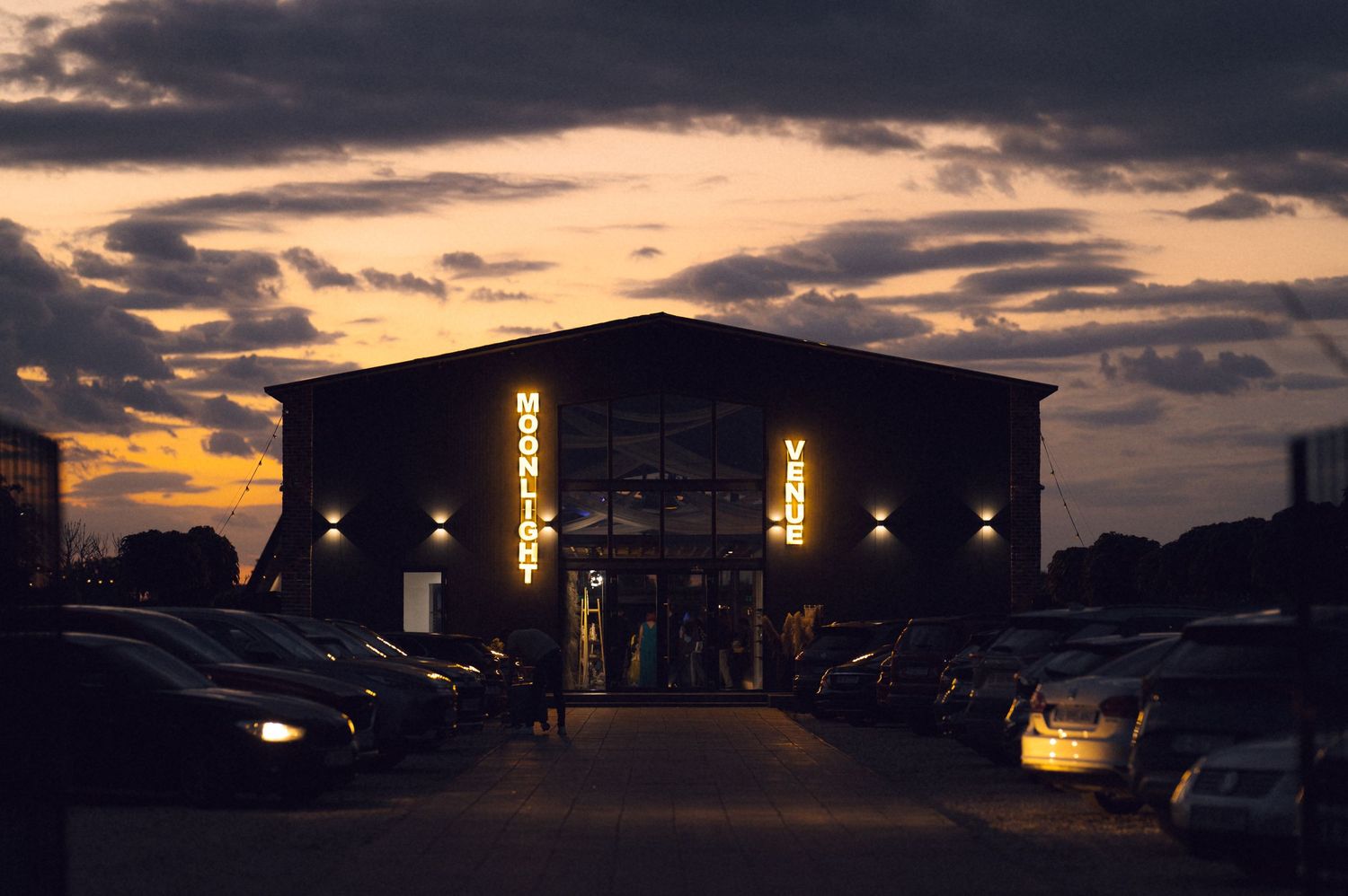 Modern venue building with illuminated signage at sunset with cars parked outside.