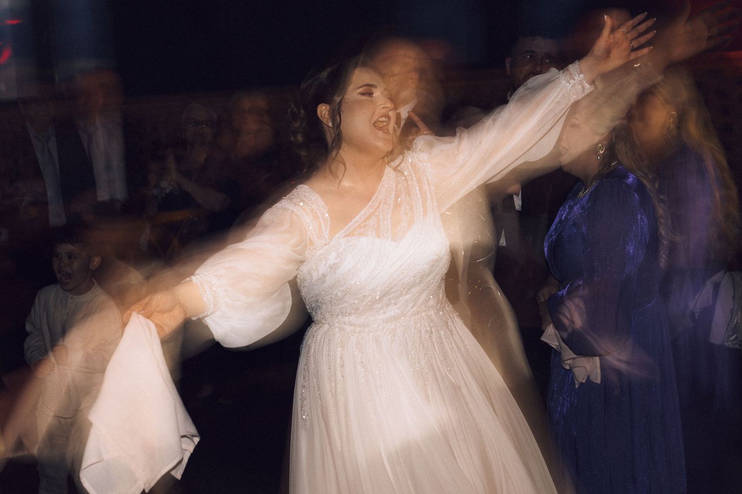 Bride in white dress dancing joyfully with arms outstretched at wedding reception with motion blur effect.