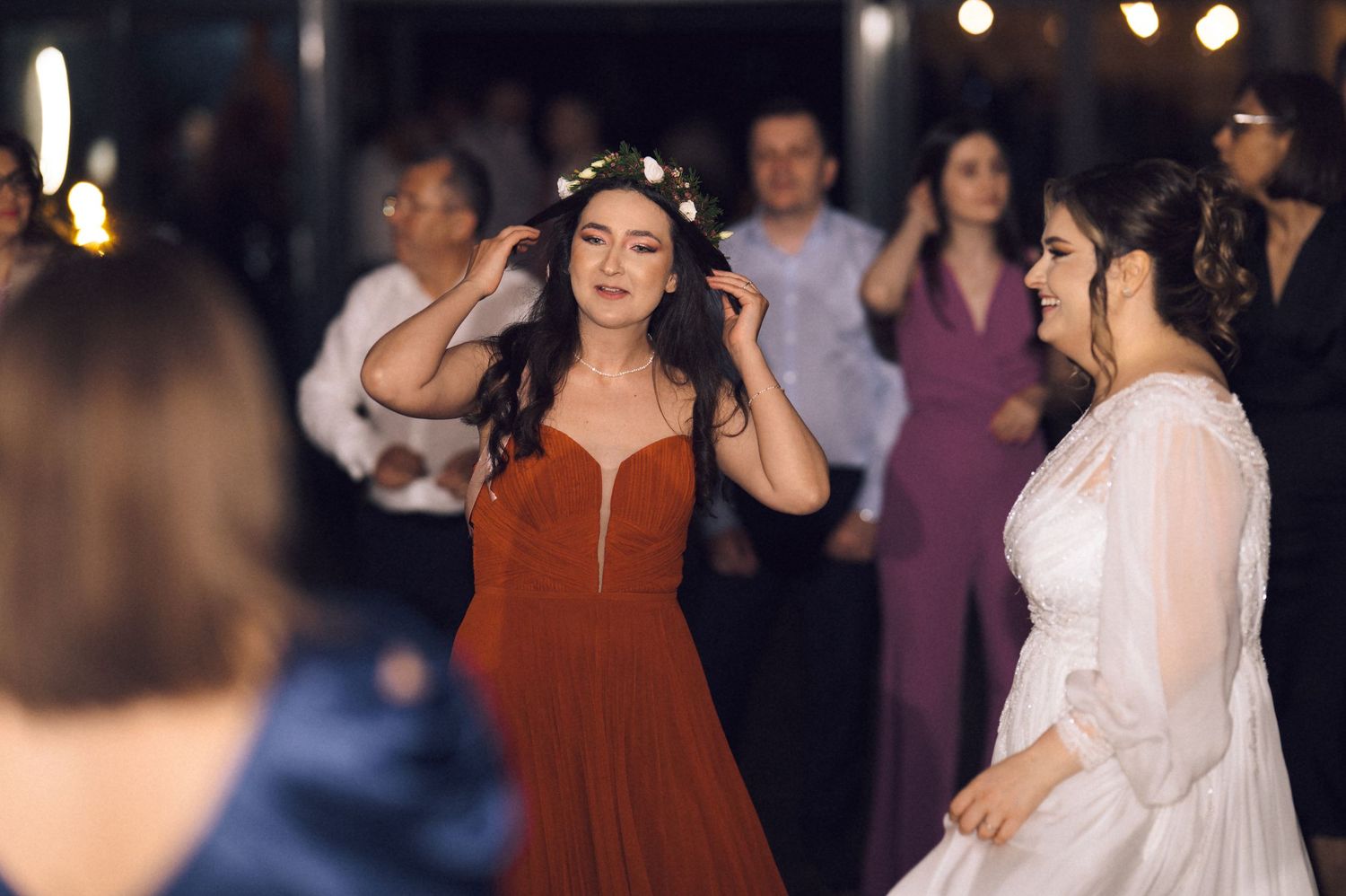 Two women dancing joyfully at a wedding reception, one in a red gown with floral crown, the other in white bridal attire.