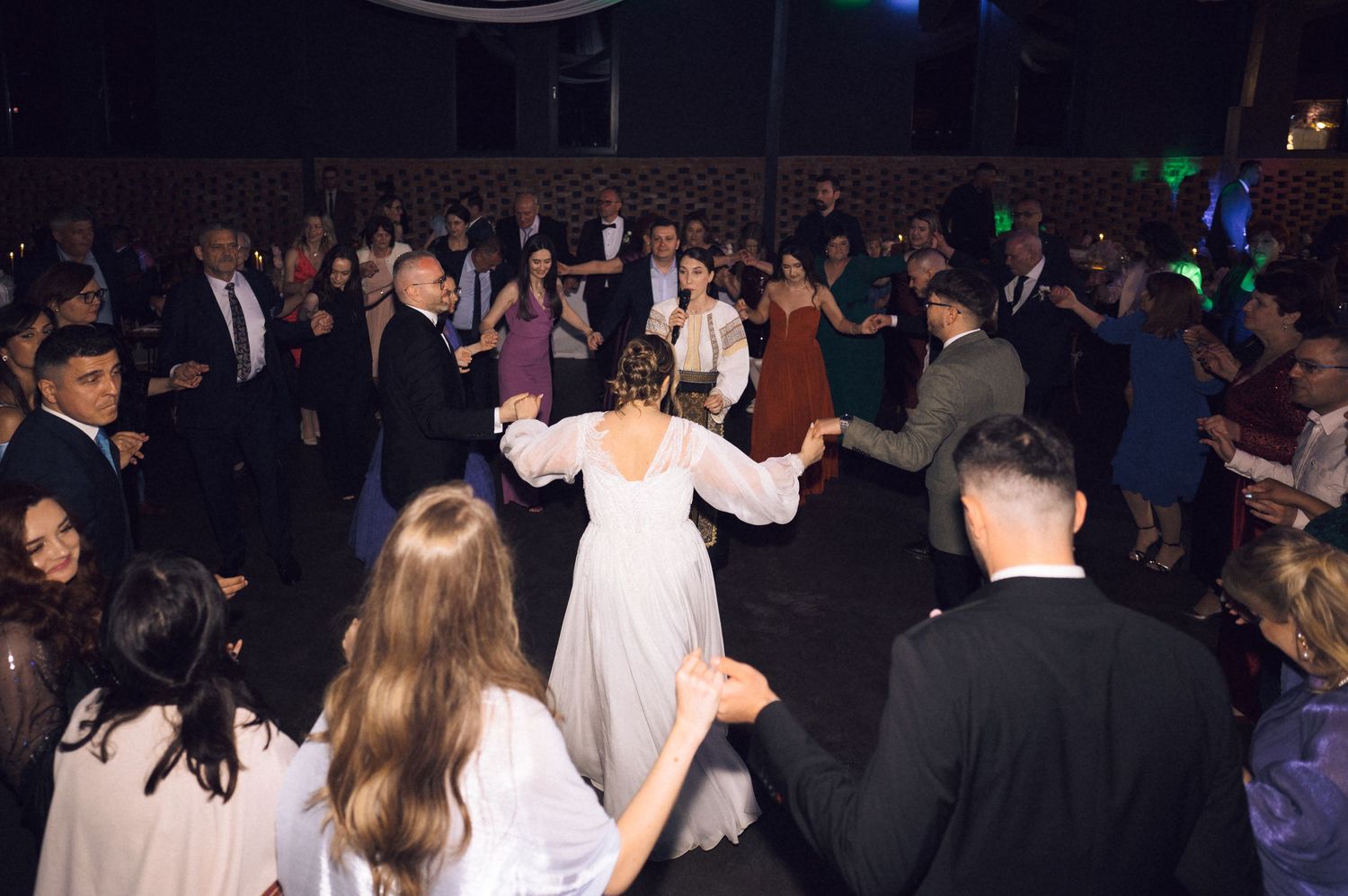 Bride in white gown dancing with guests on a crowded wedding reception dance floor at an elegant evening celebration.