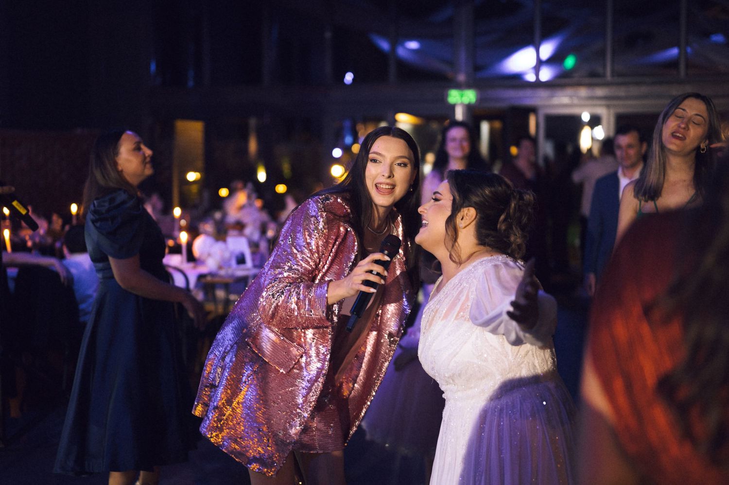 Two women laugh and dance together at a glamorous evening party, one wearing a stunning pink sequin jacket.